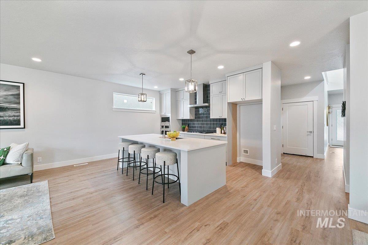 Kitchen featuring a breakfast bar, backsplash, pendant lighting, light wood finished floors, and wall chimney exhaust hood