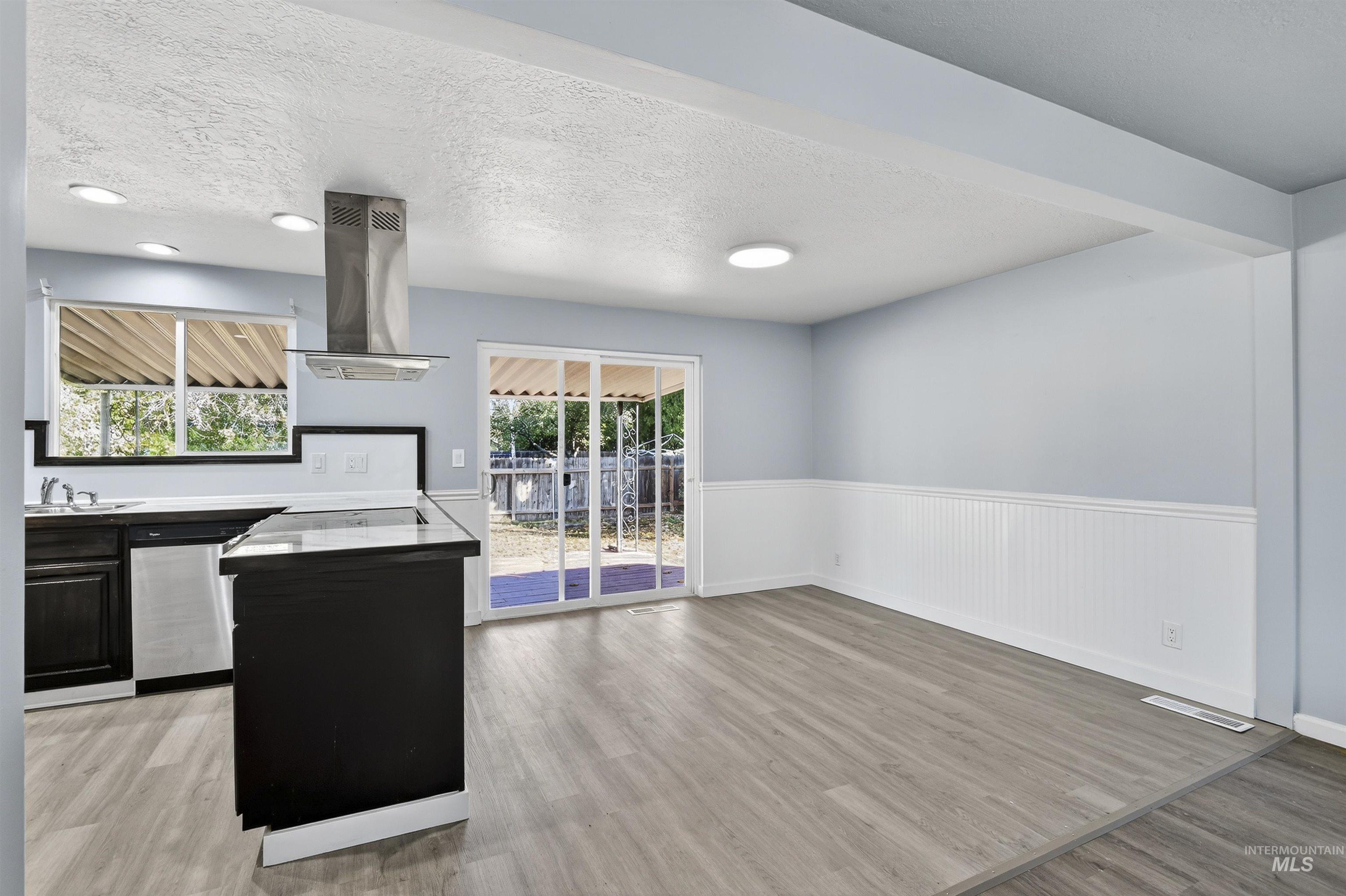 Kitchen with a textured ceiling, light wood-type flooring, island range hood, a wainscoted wall, and stainless steel dishwasher