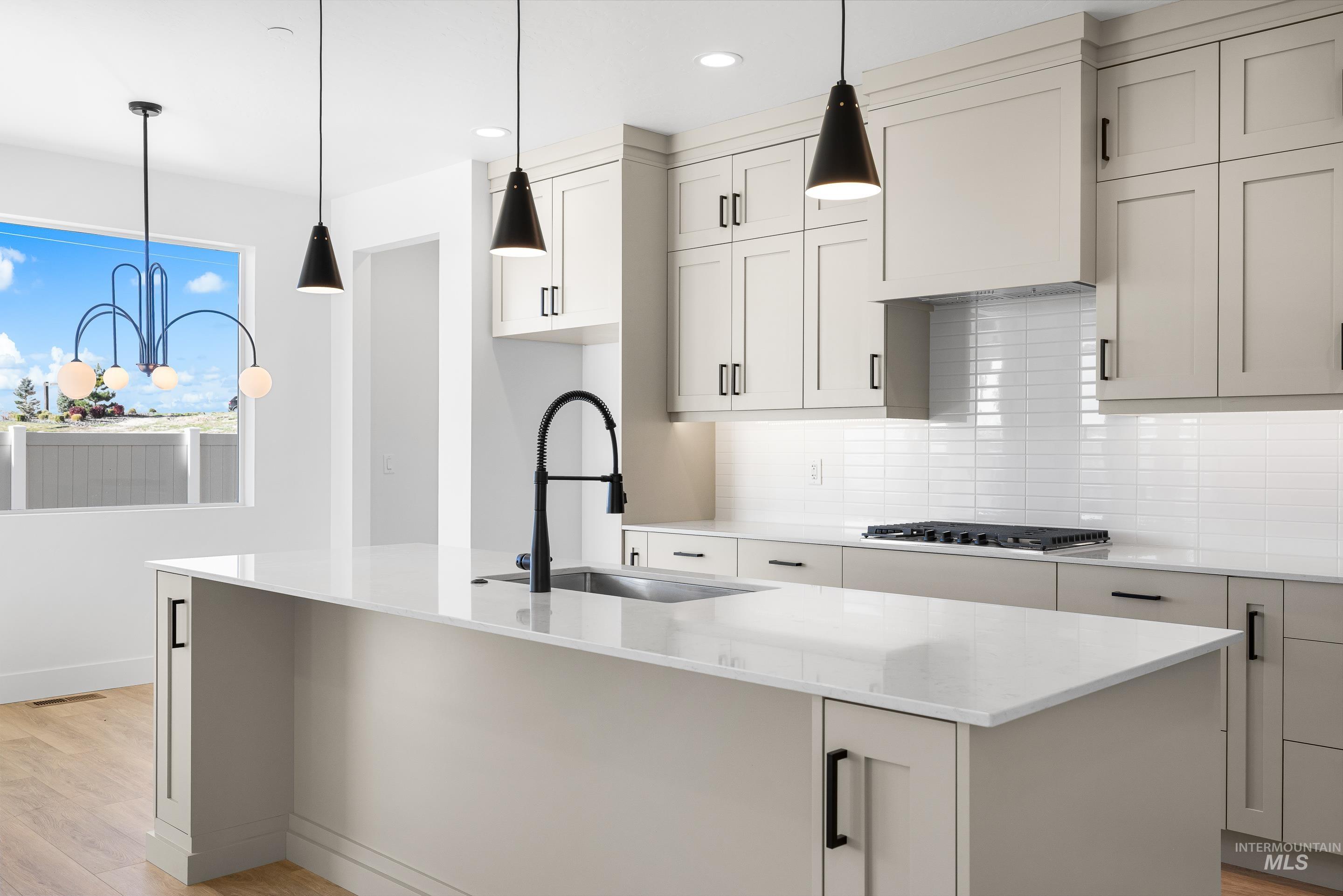 Kitchen with backsplash, light stone counters, light wood finished floors, a center island with sink, and decorative light fixtures