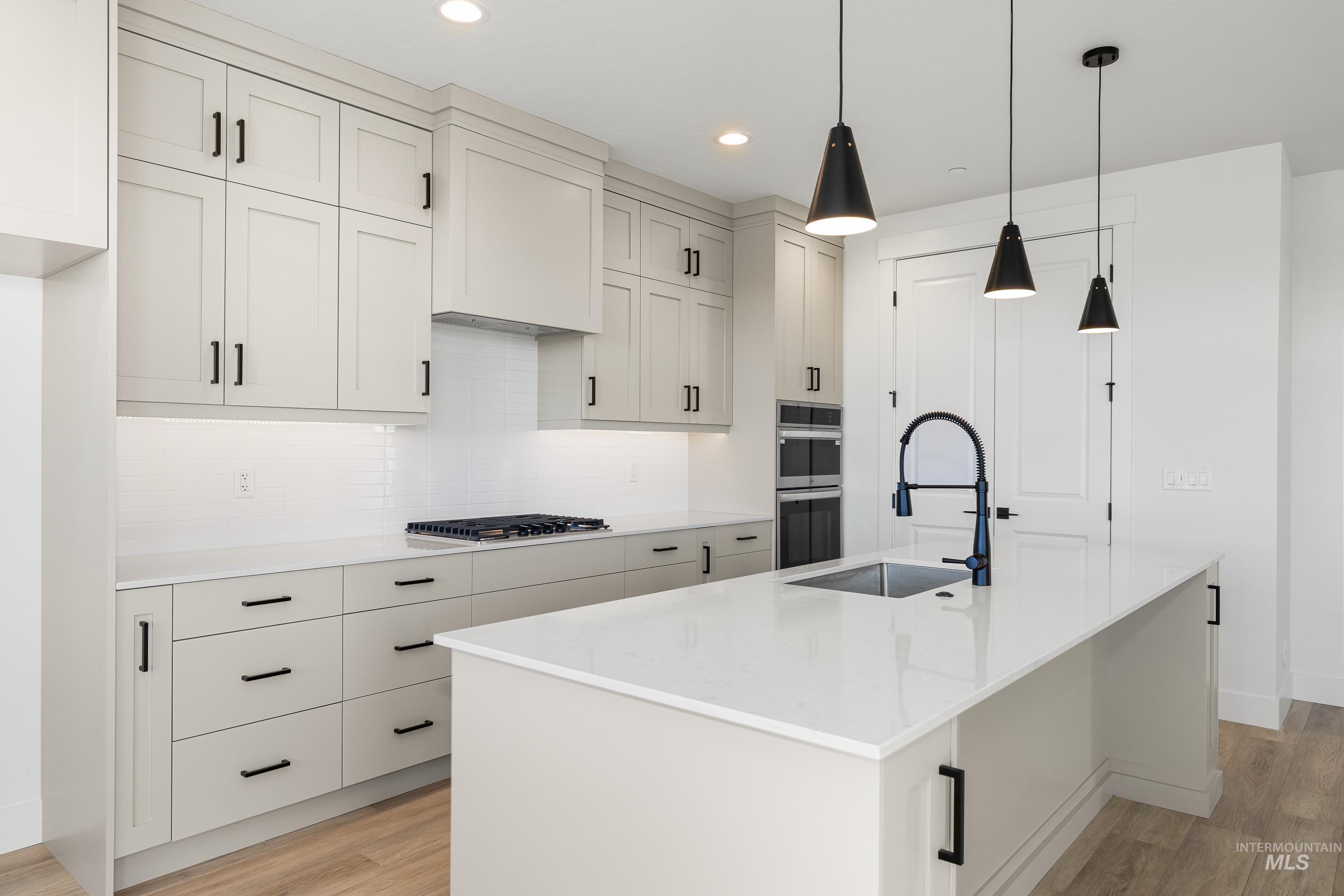 Kitchen with light wood-type flooring, a center island with sink, light stone counters, backsplash, and decorative light fixtures