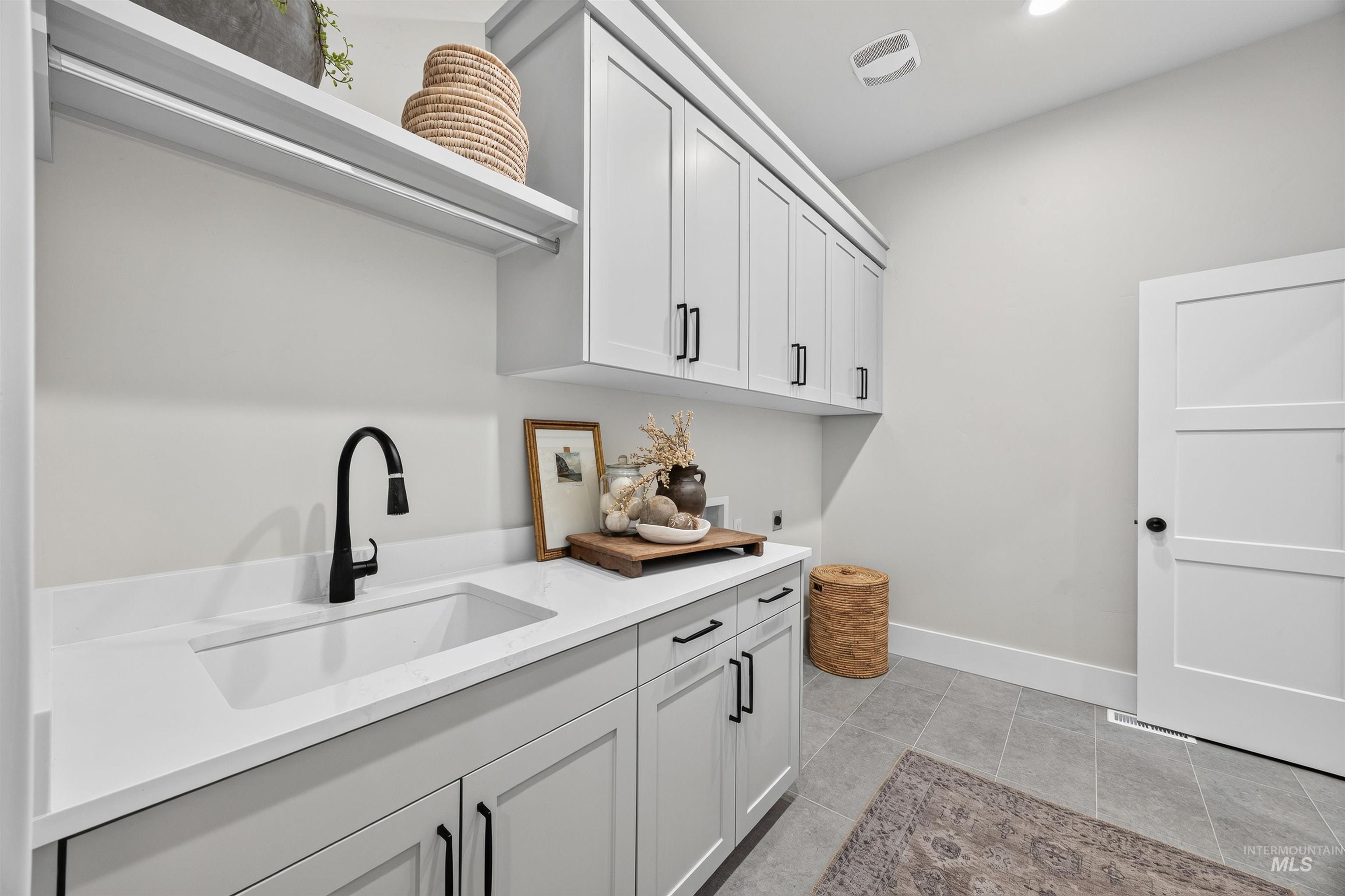 Laundry room featuring light gray cabinetry with black hardware, white solid surface countertops, an integrated sink with a matte black faucet, and gray tile flooring