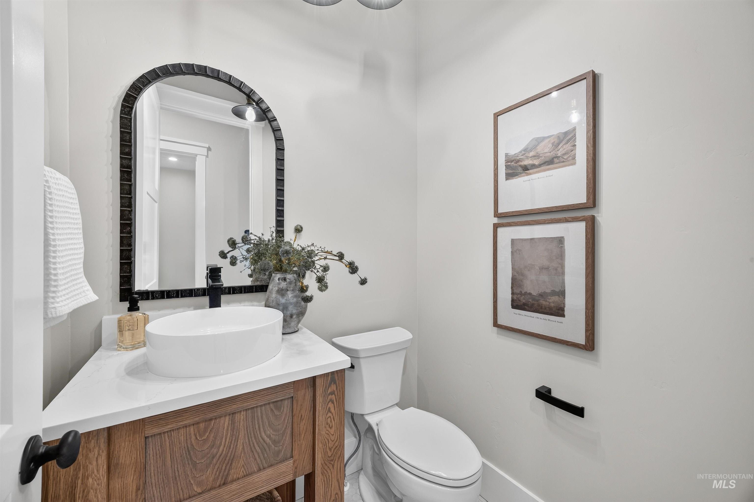 Powder room featuring a wood-finish vanity with a white countertop, an oval vessel sink, and an arched framed mirror