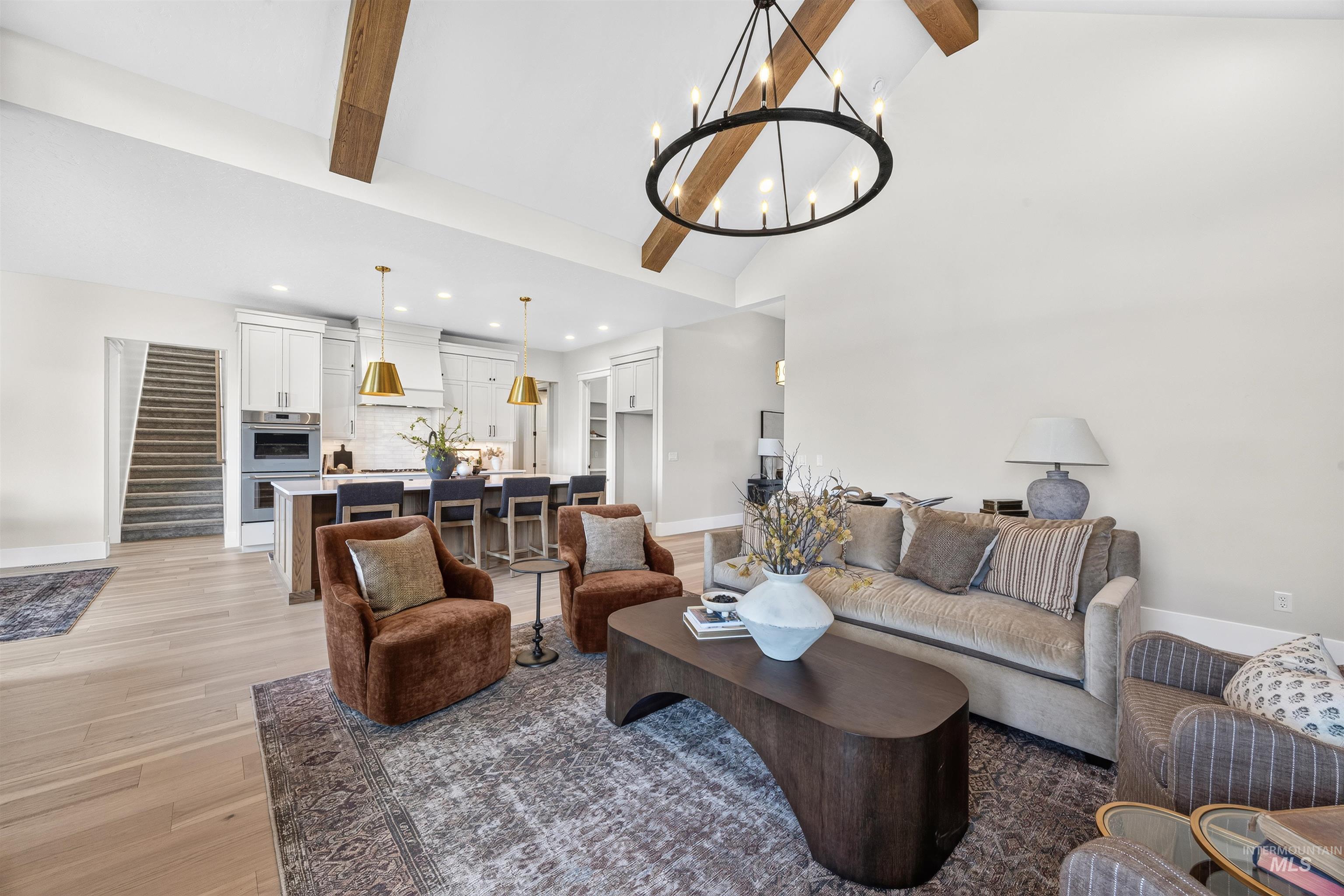 Spacious open-concept interior featuring light wood-finish flooring, vaulted ceilings with exposed wood beams, and a large circular chandelier