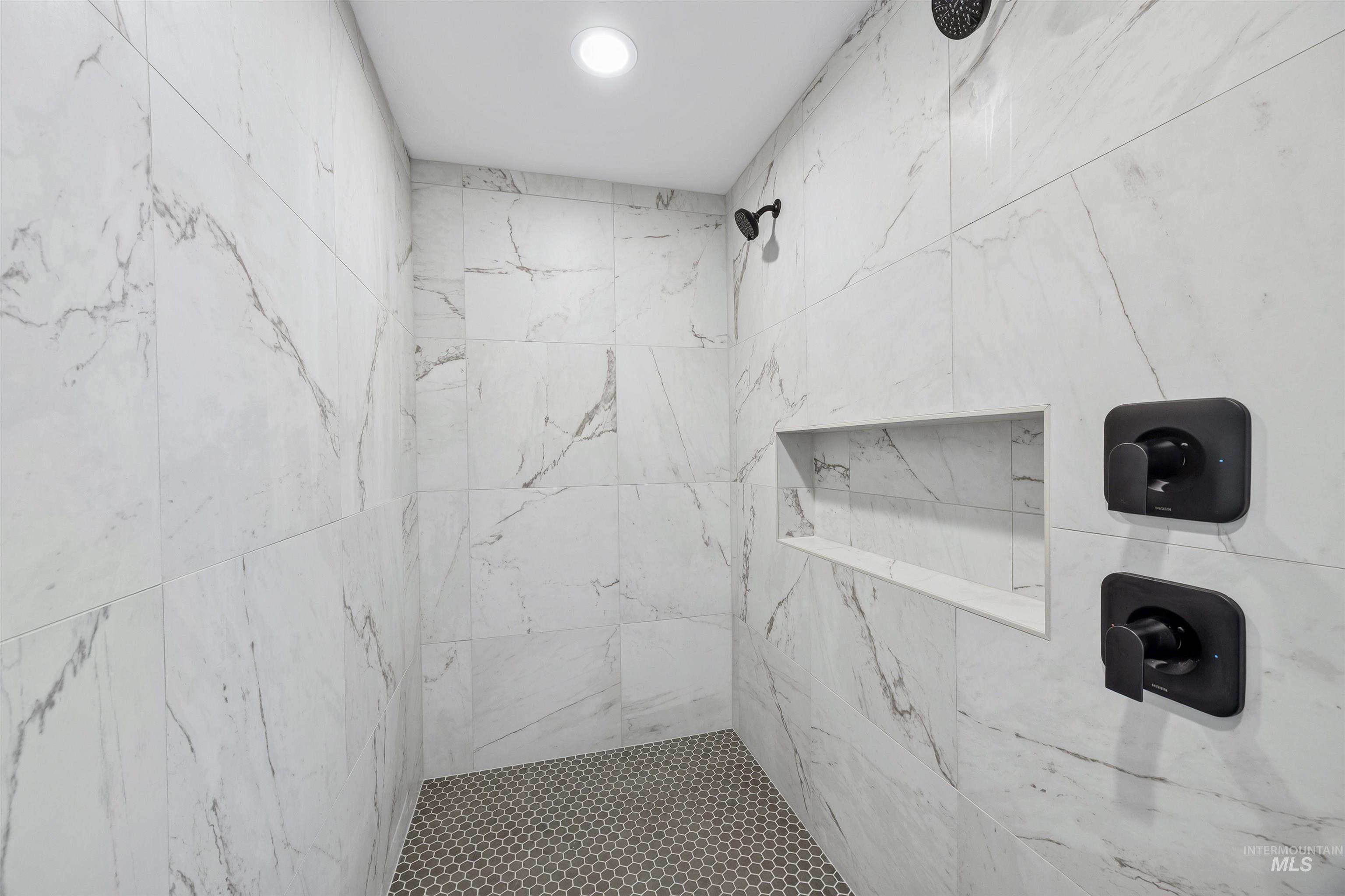 Spacious shower featuring large format marble-look wall tiles, hexagonal floor tiles, a built-in shower niche, and matte black fixtures including a rain shower head and separate shower head