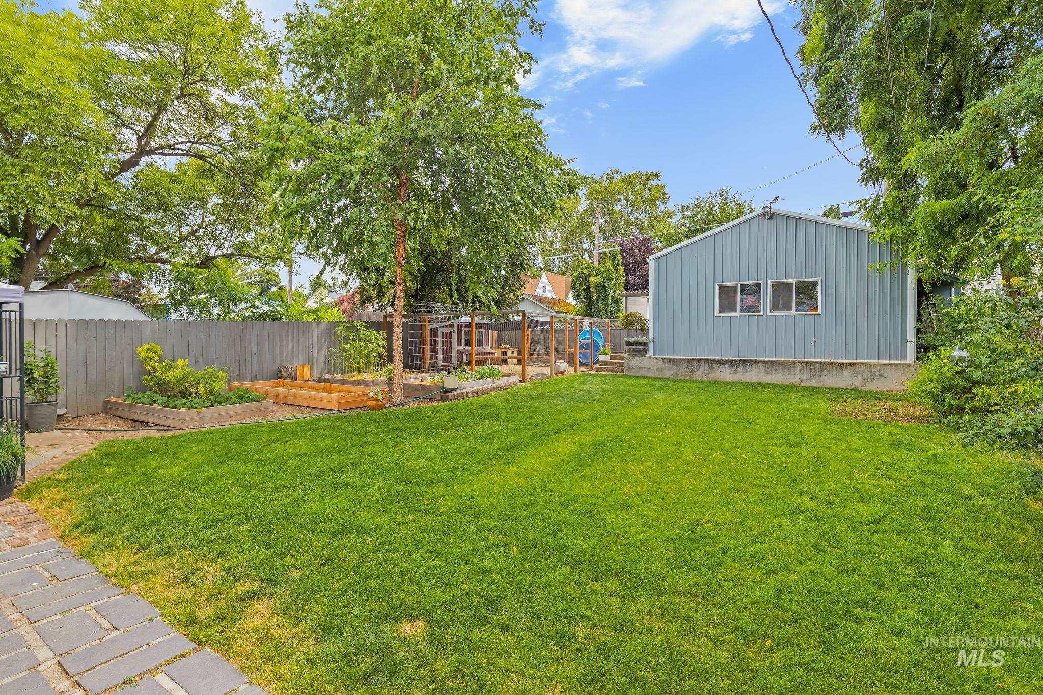 Spacious backyard featuring a large green lawn, mature trees, a privacy fence, raised garden beds, and a detached structure with three windows