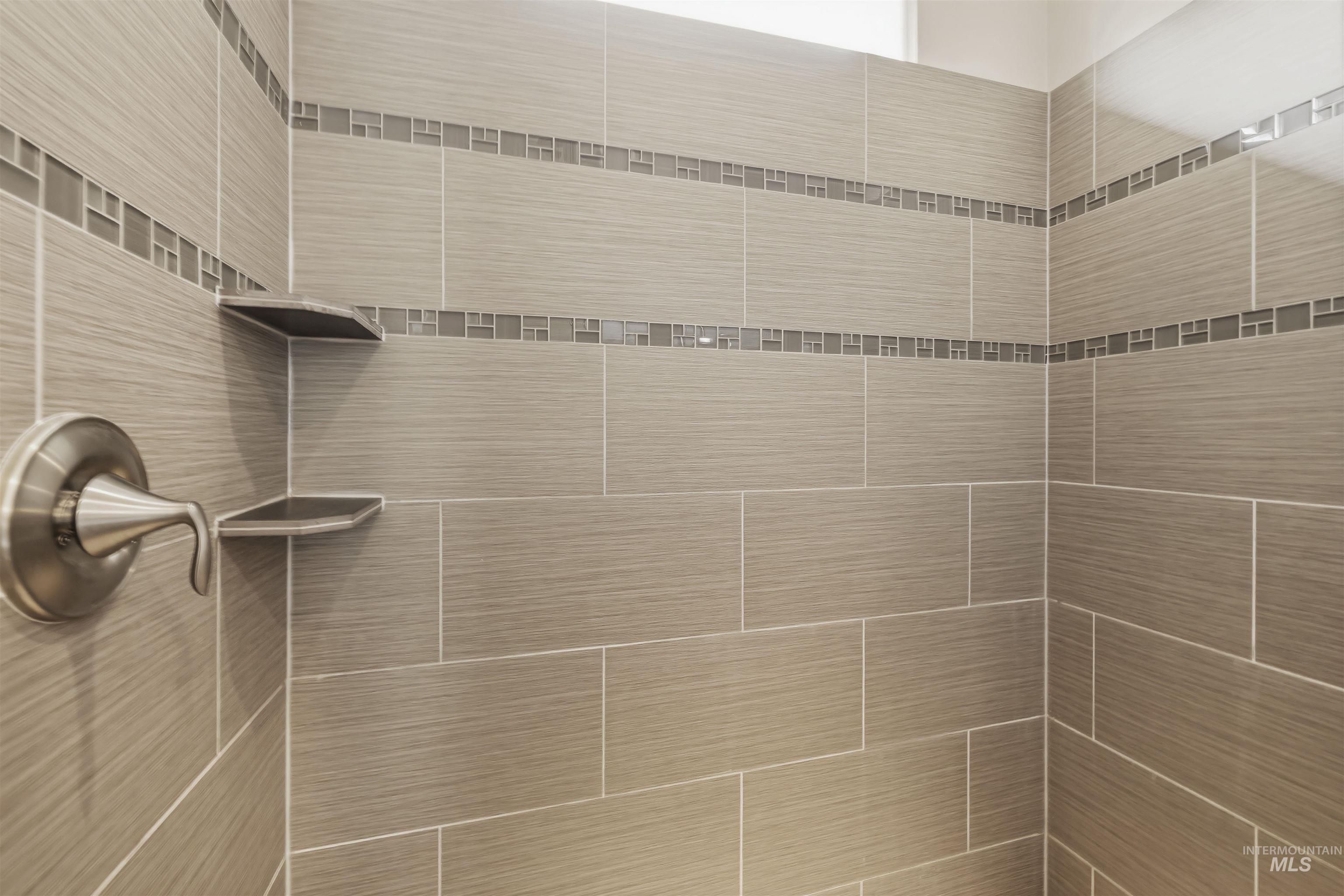 Shower enclosure featuring large format grey tile with linear texture, decorative mosaic accent strips, brushed nickel fixture, and two built-in corner shelves