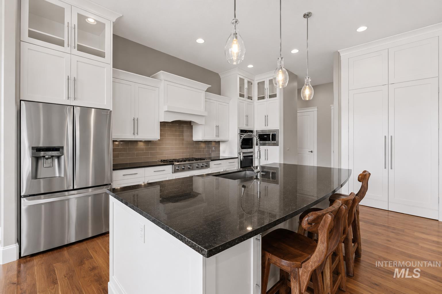 Contemporary kitchen featuring a large central island with a dark countertop and integrated sink, white cabinetry with glass-front uppers, stainless steel appliances, and wood-finish flooring