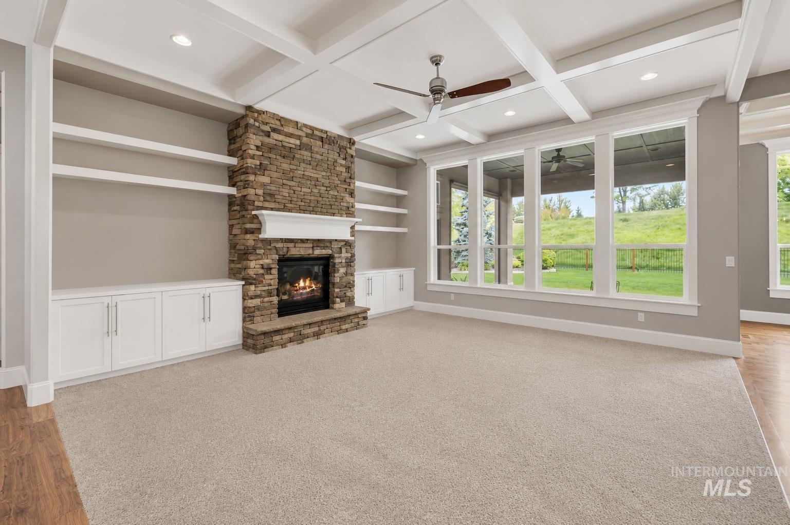 Spacious living area featuring a stacked stone fireplace with white mantle, custom built-in cabinetry and shelving, a coffered ceiling with recessed lighting and ceiling fan, and large windows revealing an outdoor landscape
