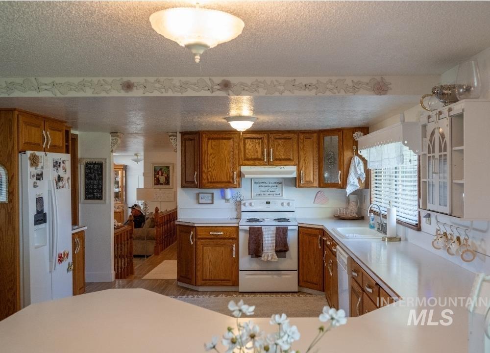 Kitchen with brown cabinets, white appliances, light countertops, a textured ceiling, and under cabinet range hood