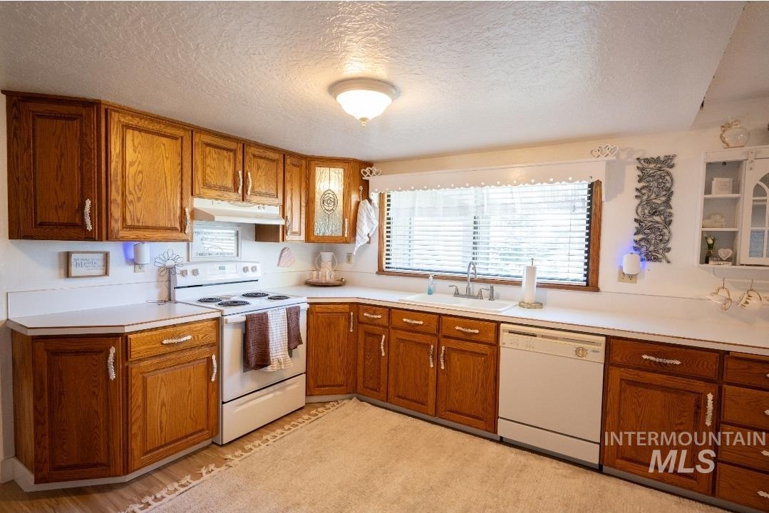 Kitchen with brown cabinets, white appliances, a textured ceiling, light countertops, and under cabinet range hood