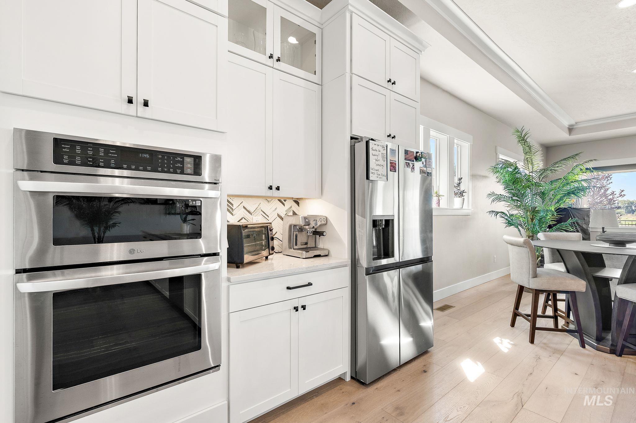Kitchen with stainless steel appliances, white cabinetry, light wood-style floors, glass insert cabinets, and decorative backsplash