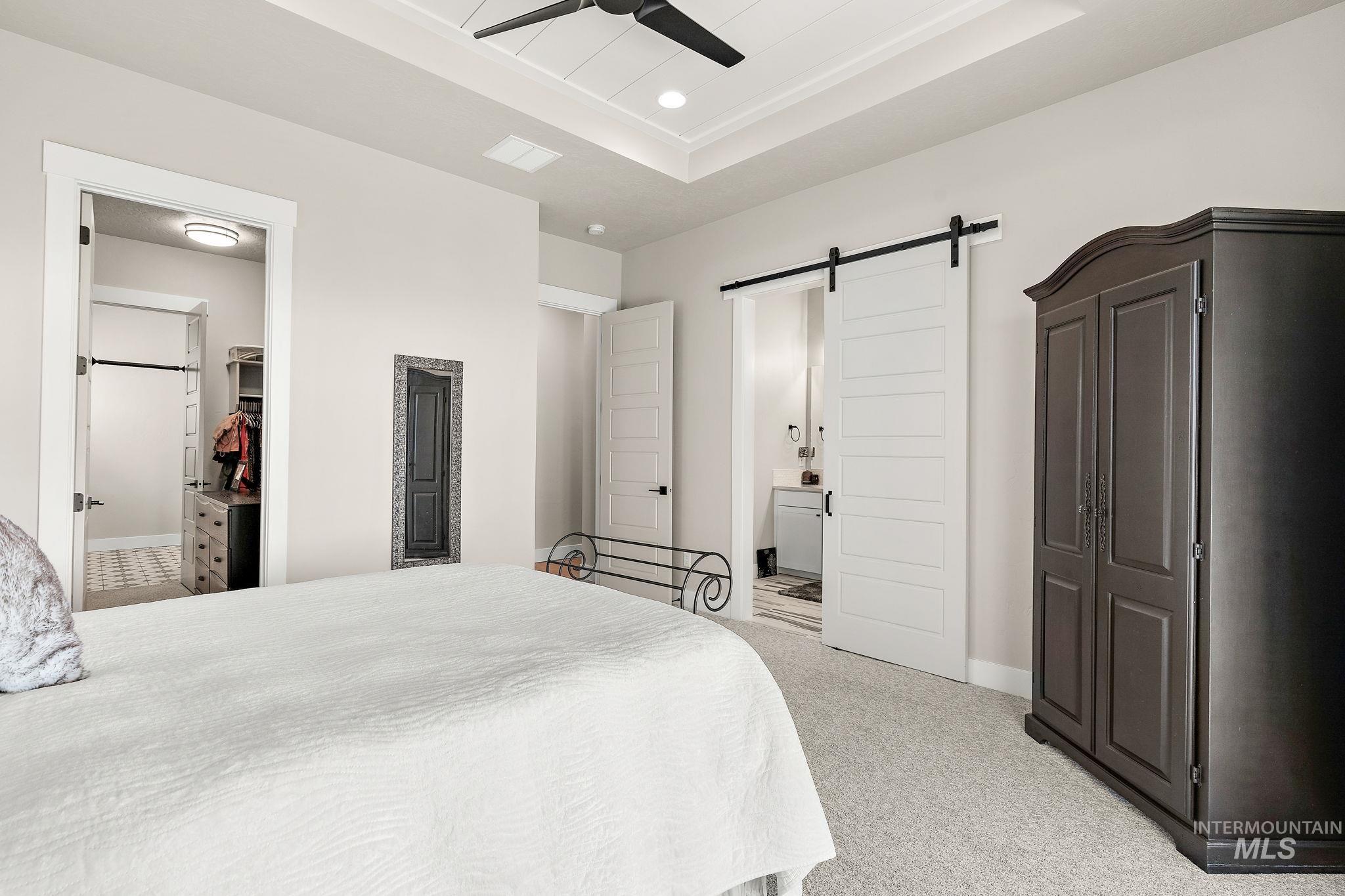 Bedroom with a barn door, ensuite bath, a raised ceiling, light carpet, and a ceiling fan