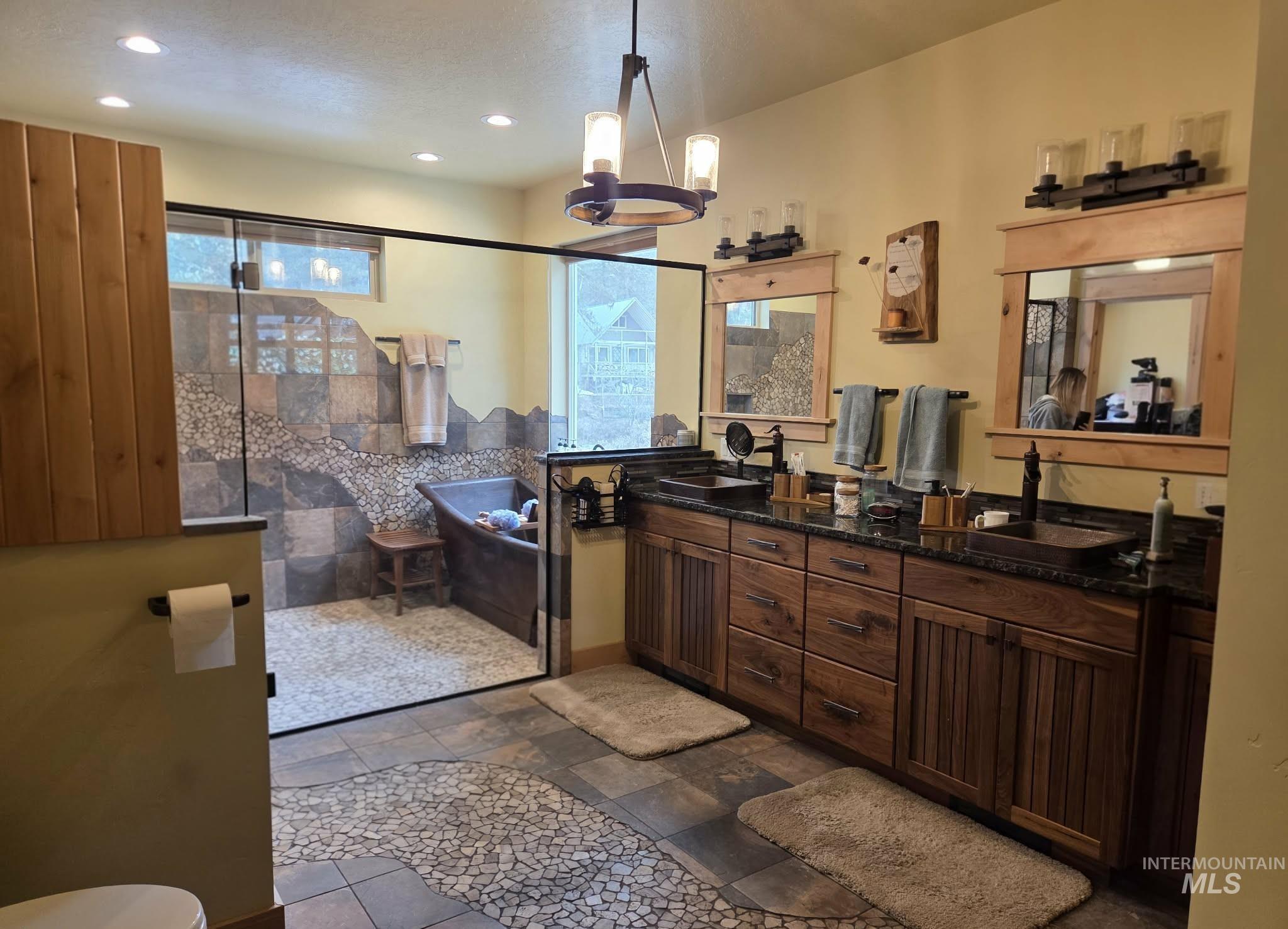 Full bath with double vanity, a stall shower, a chandelier, stone tile floors, and a bathtub