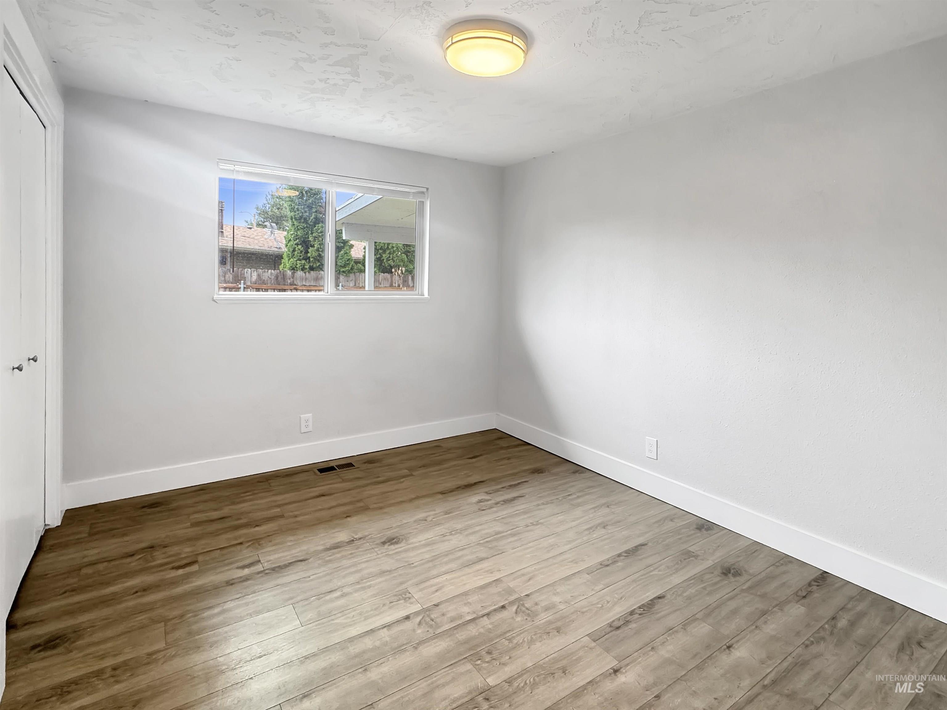 Room featuring wood-finish flooring, white baseboards, a window with exterior views, a ceiling-mounted light fixture, and white bi-fold closet doors