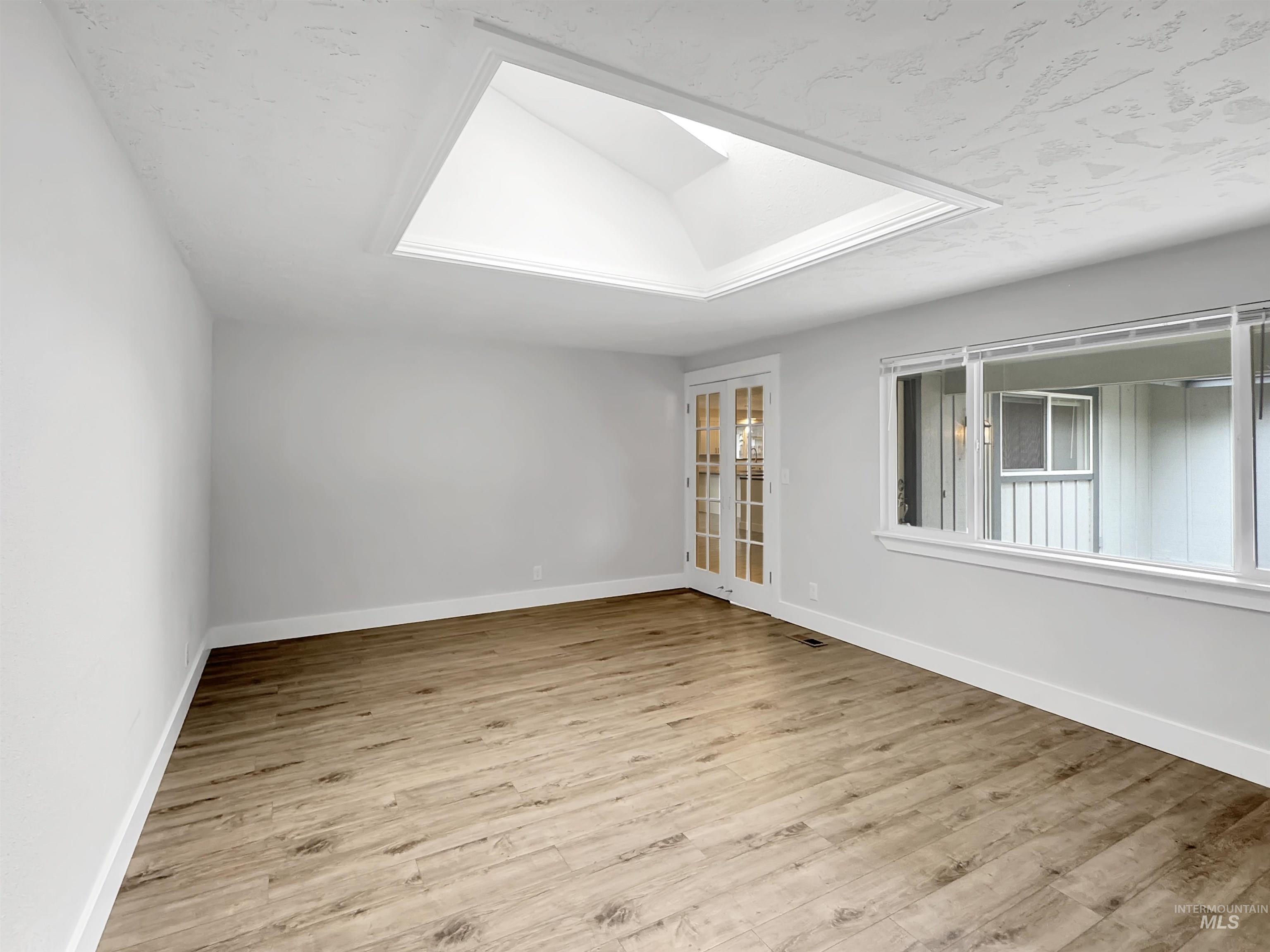 Spacious room featuring a skylight, wood-finish flooring, white walls, large windows, and double French doors
