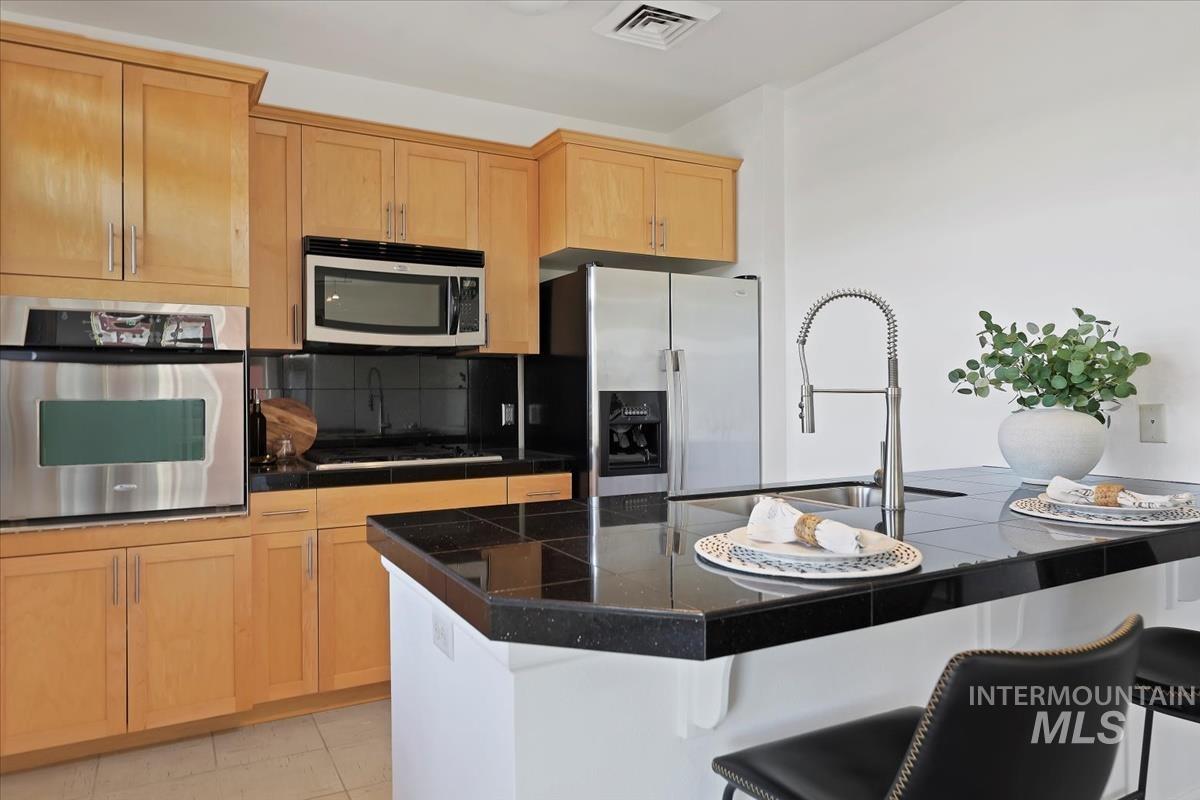 Kitchen featuring appliances with stainless steel finishes, a breakfast bar, tile countertops, a kitchen island with sink, and light brown cabinetry