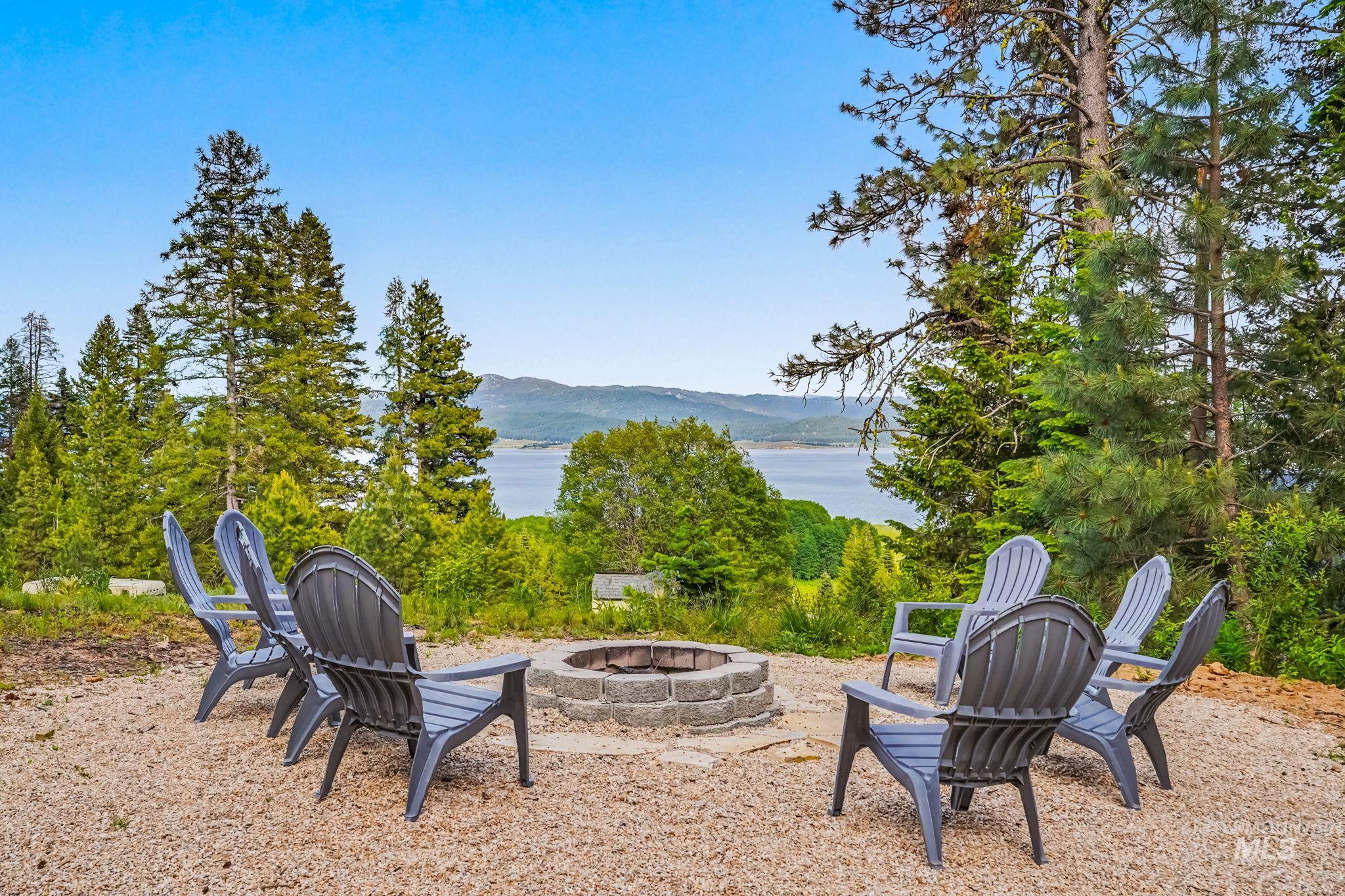 Gravel fire pit area featuring a stone block fire pit surround, surrounded by mature trees, with distant water scenery and mountain backdrop
