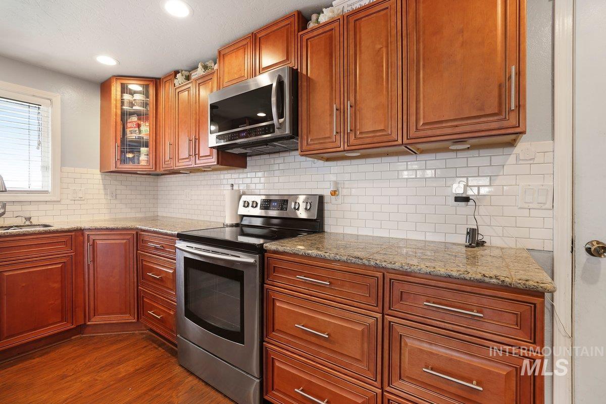 Kitchen featuring stainless steel appliances, dark wood-style flooring, glass insert cabinets, tasteful backsplash, and brown cabinetry