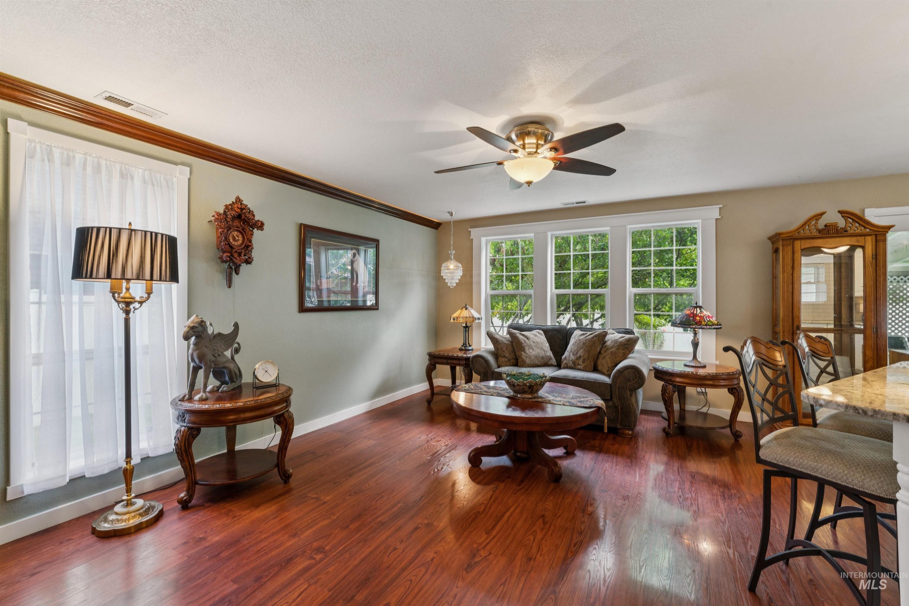 Spacious living area featuring polished wood-finish flooring, a ceiling fan with integrated lighting, multiple windows with white trim, and crown molding