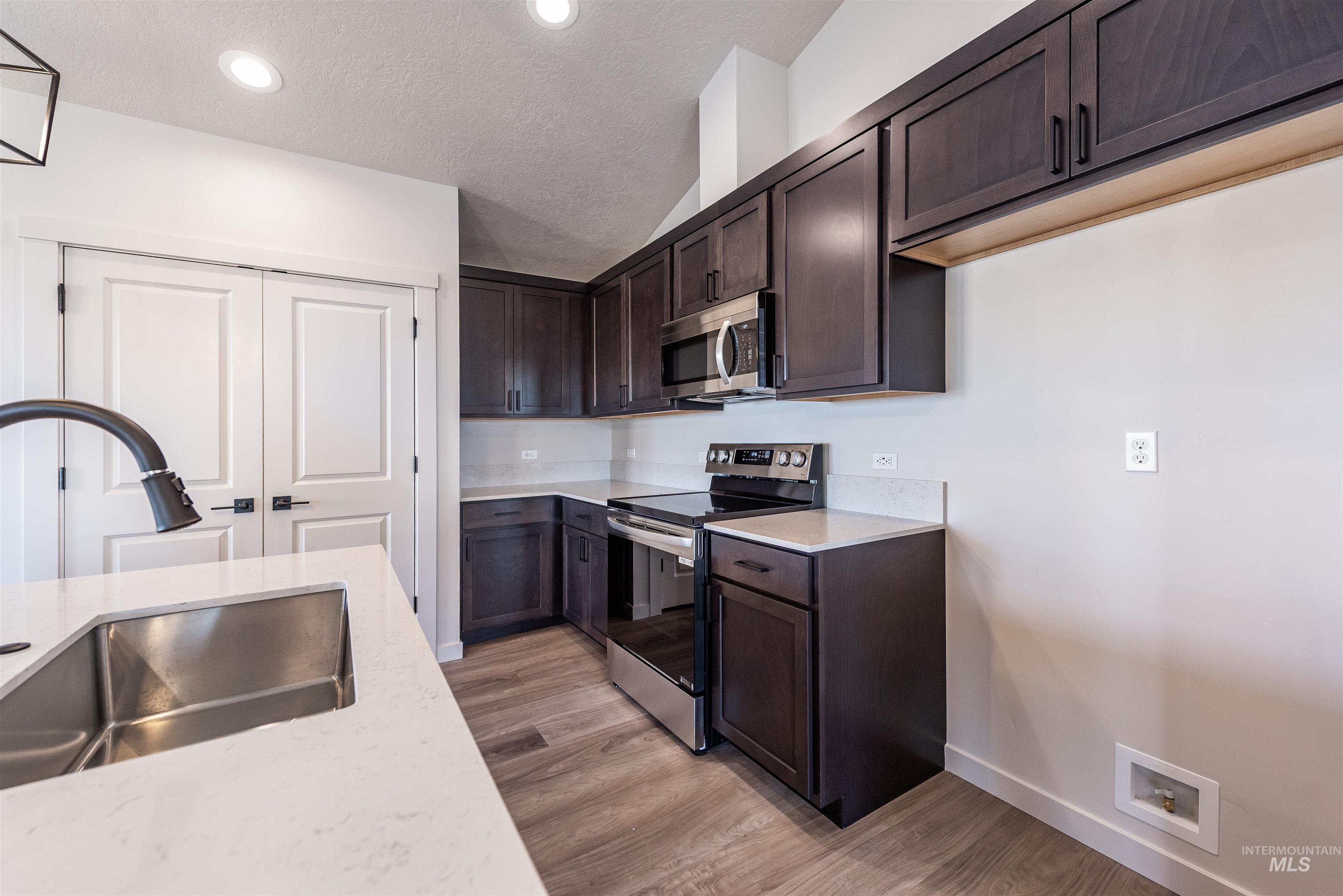 Kitchen featuring dark brown cabinets, appliances with stainless steel finishes, a textured ceiling, light wood-type flooring, and light stone countertops
