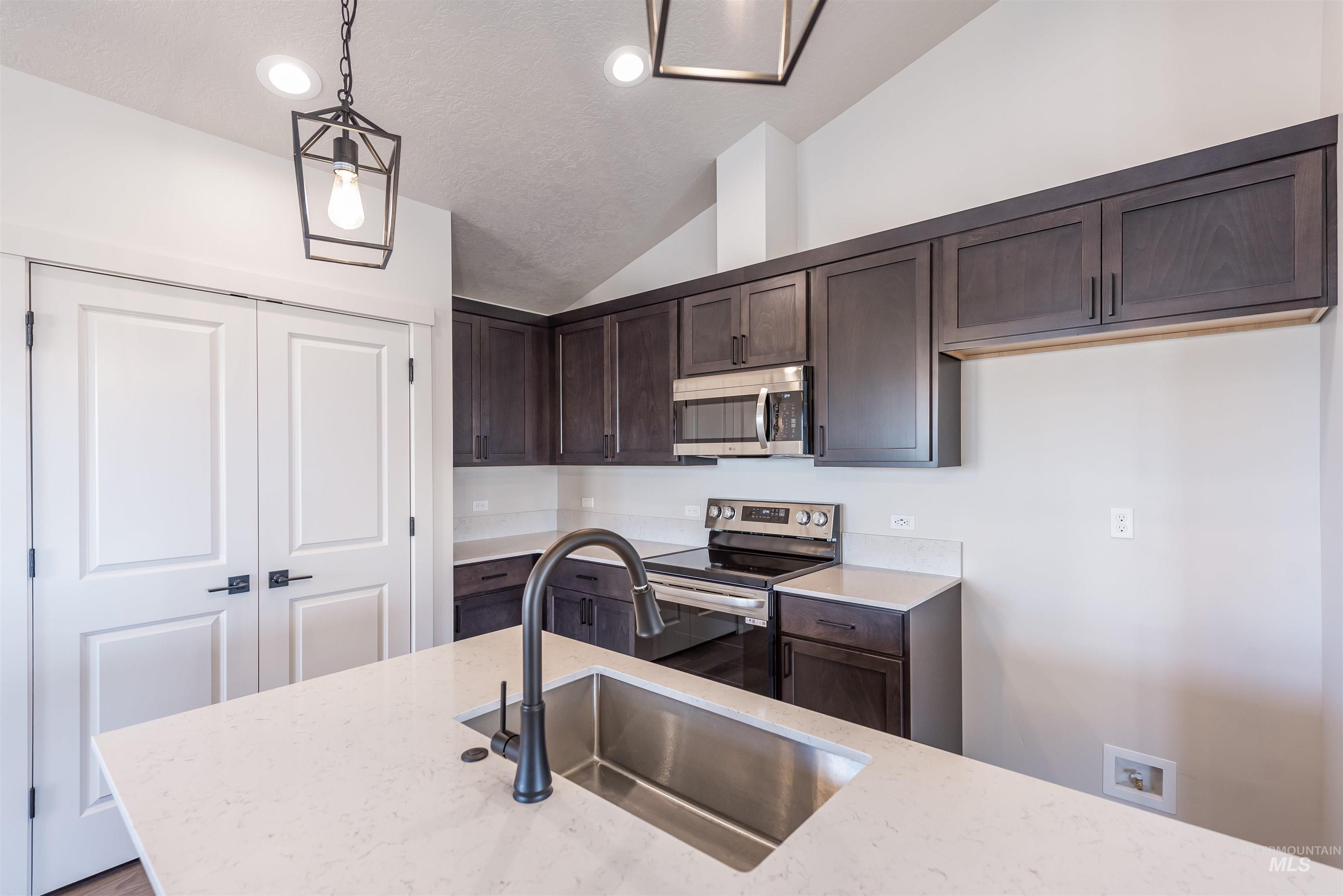 Kitchen with dark brown cabinetry, appliances with stainless steel finishes, decorative light fixtures, vaulted ceiling, and light stone countertops