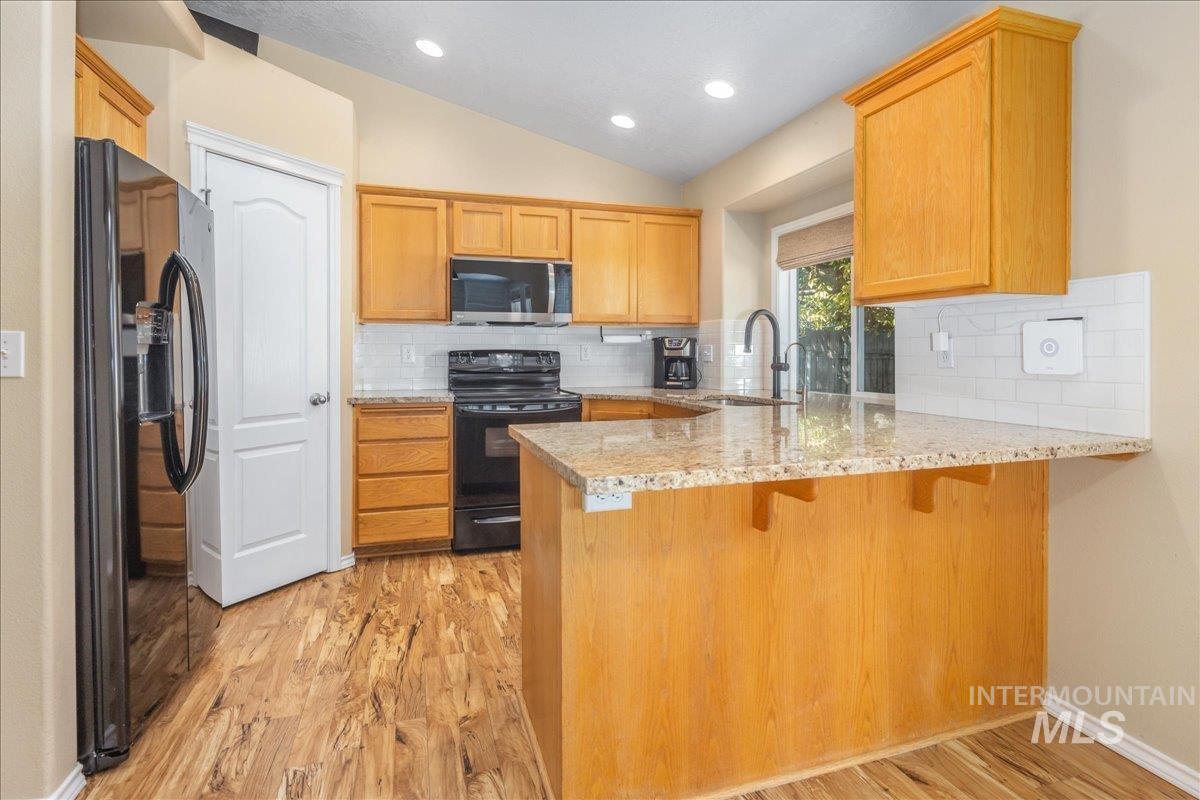Kitchen featuring a peninsula, backsplash, black appliances, light wood-type flooring, and light stone counters