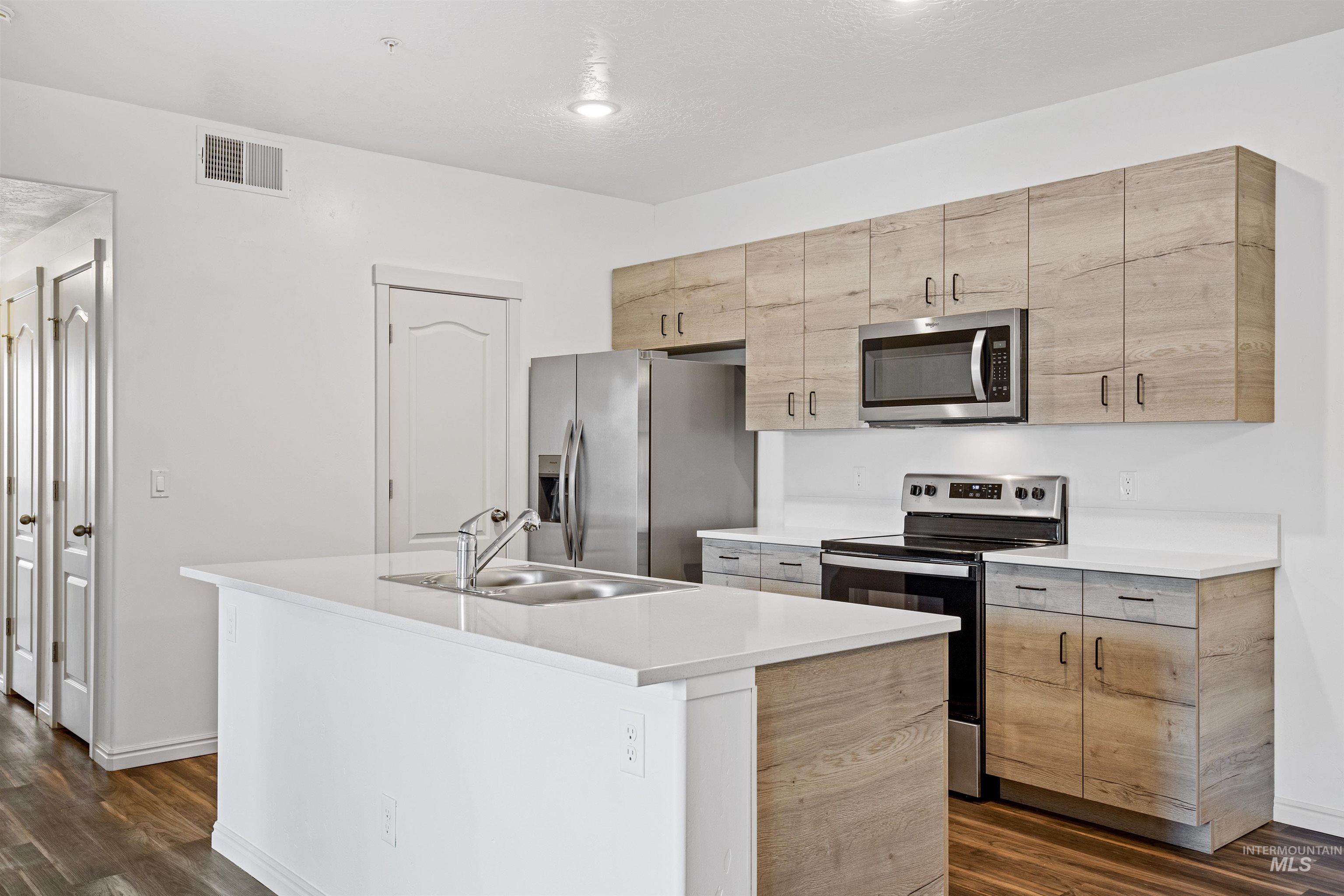 Kitchen with appliances with stainless steel finishes, dark wood-style floors, an island with sink, light countertops, and light brown cabinetry