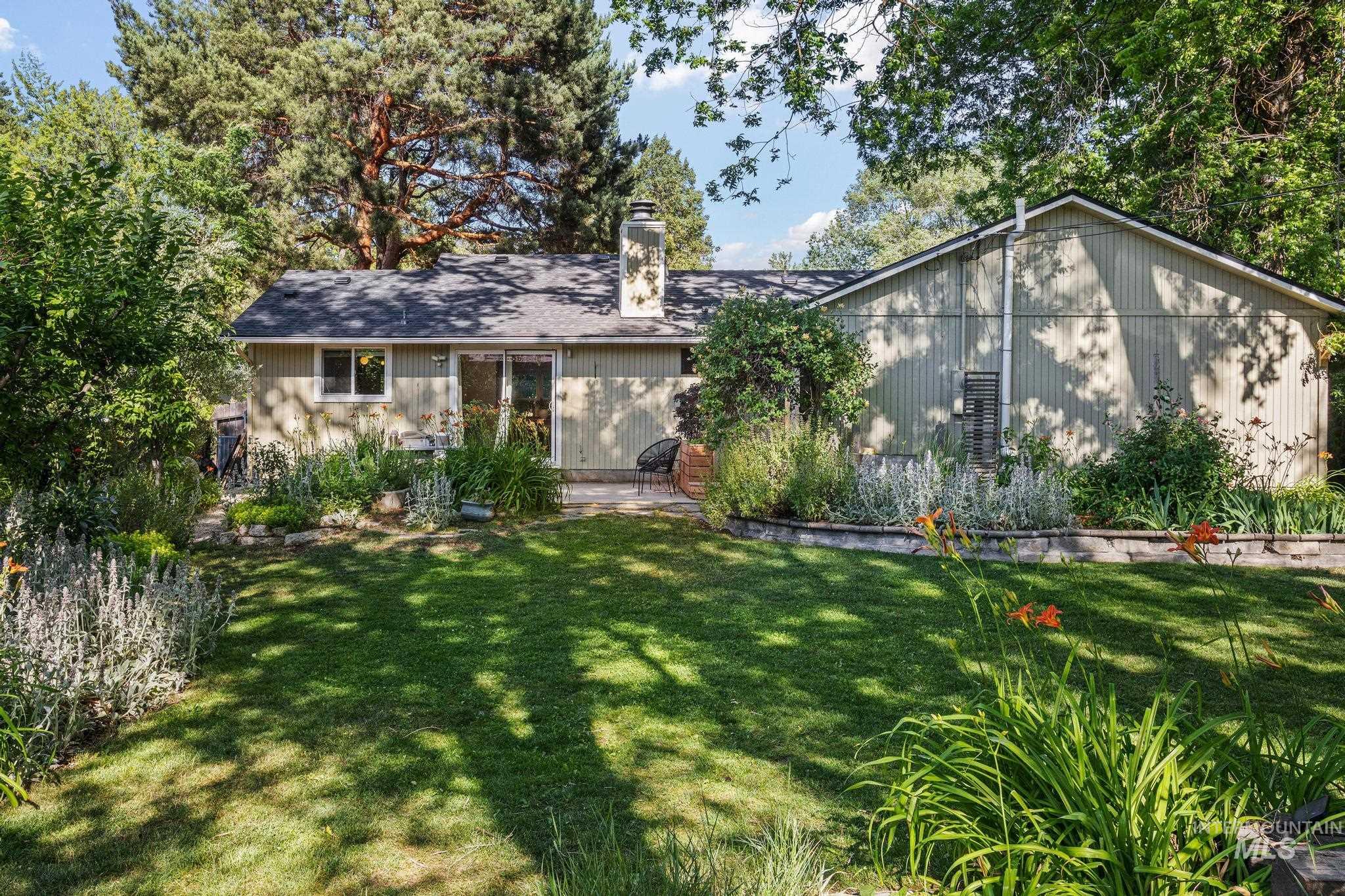 Expansive lawn area with mature perimeter trees, established garden beds, a sliding glass door, and a permanent patio space