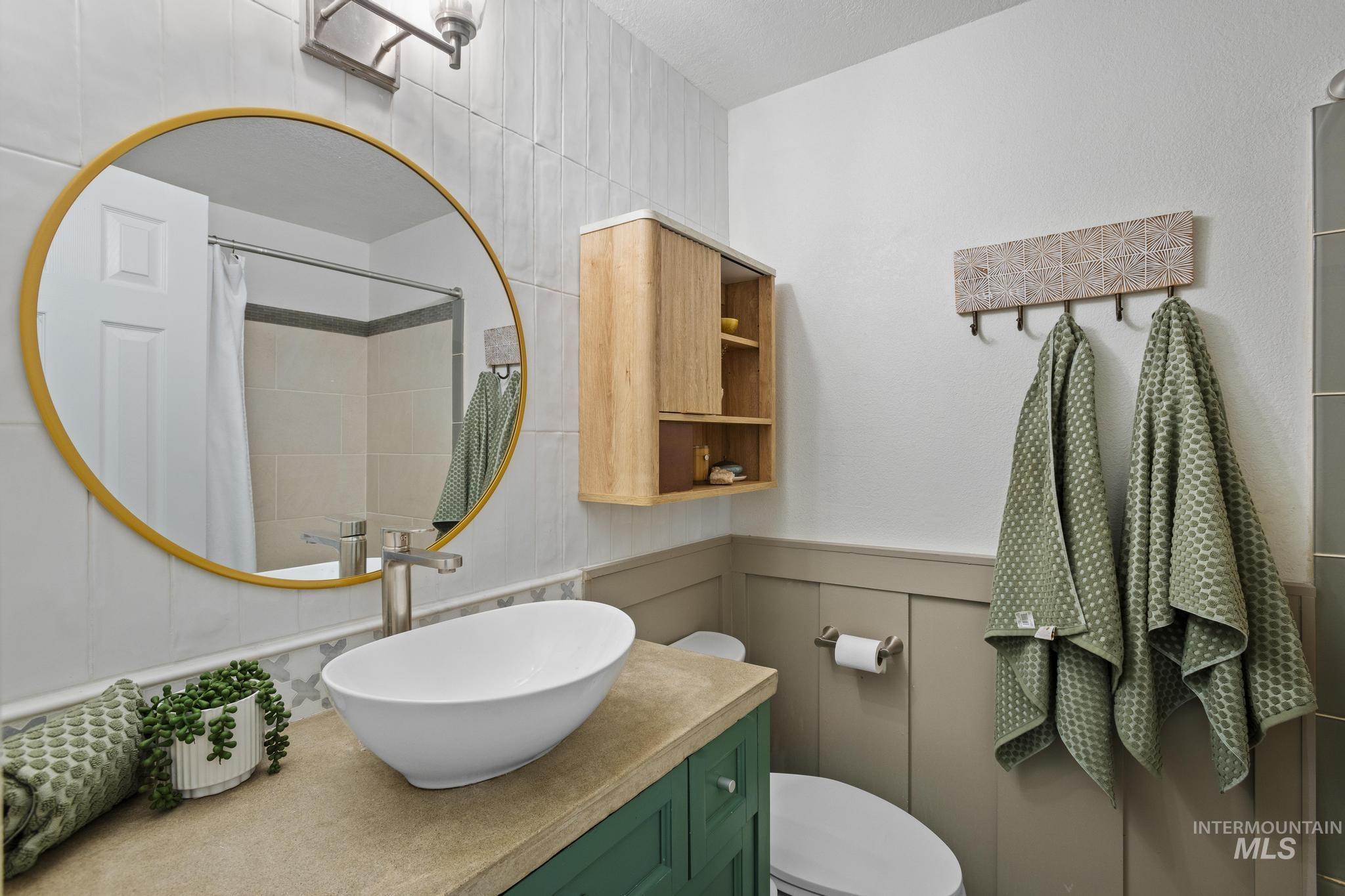 Bathroom featuring a vessel sink with a brushed nickel faucet, a gold-framed circular mirror, a wood-finish wall cabinet, wainscoting, and white wall tile