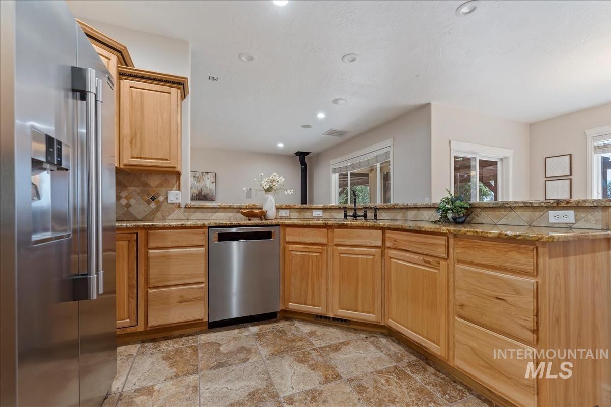Kitchen featuring natural wood cabinetry, stone-finish tile flooring, a stainless steel refrigerator with an ice and water dispenser, a stainless steel dishwasher, and stone countertops with a patterned tile backsplash