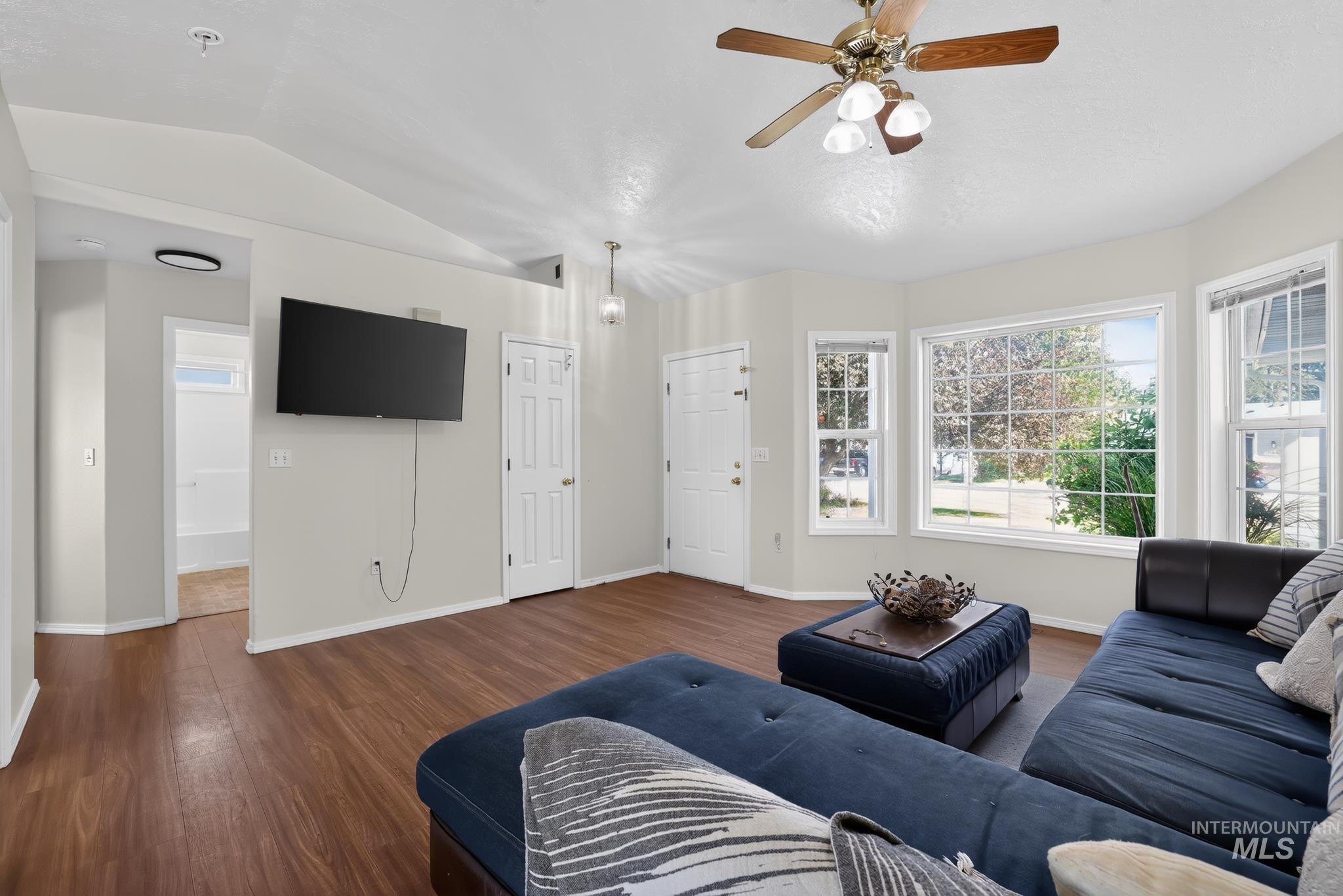Spacious living area featuring wood-finish flooring, a ceiling fan with integrated lighting, and multiple windows