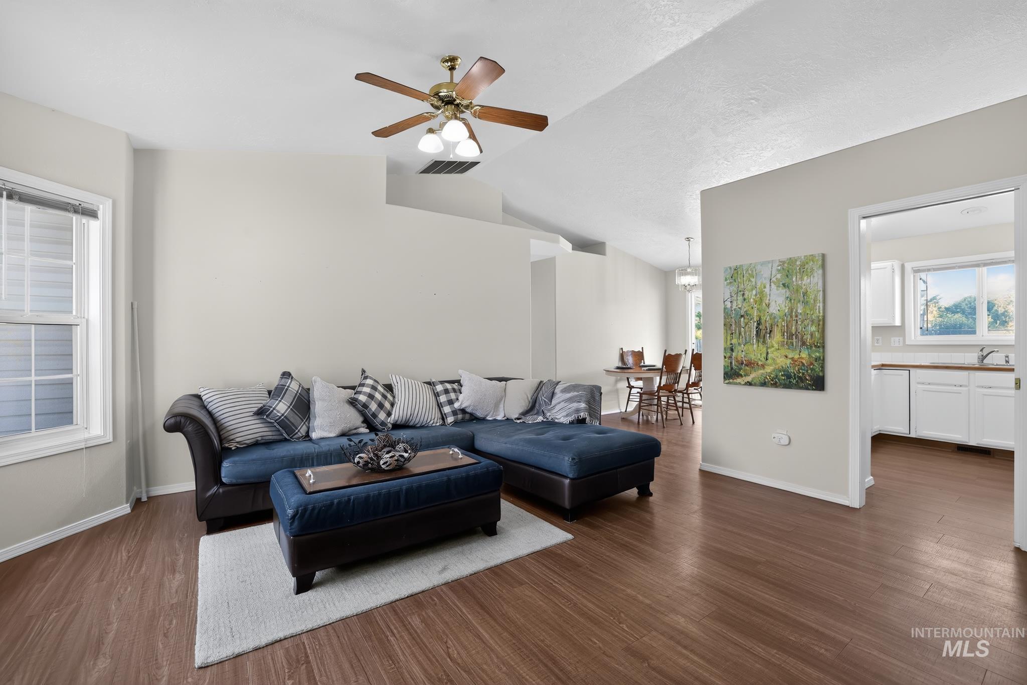 Vaulted ceiling with a ceiling fan, wood-finish flooring, ample natural light from a double-hung window, and a clear sightline to the dining area and kitchen