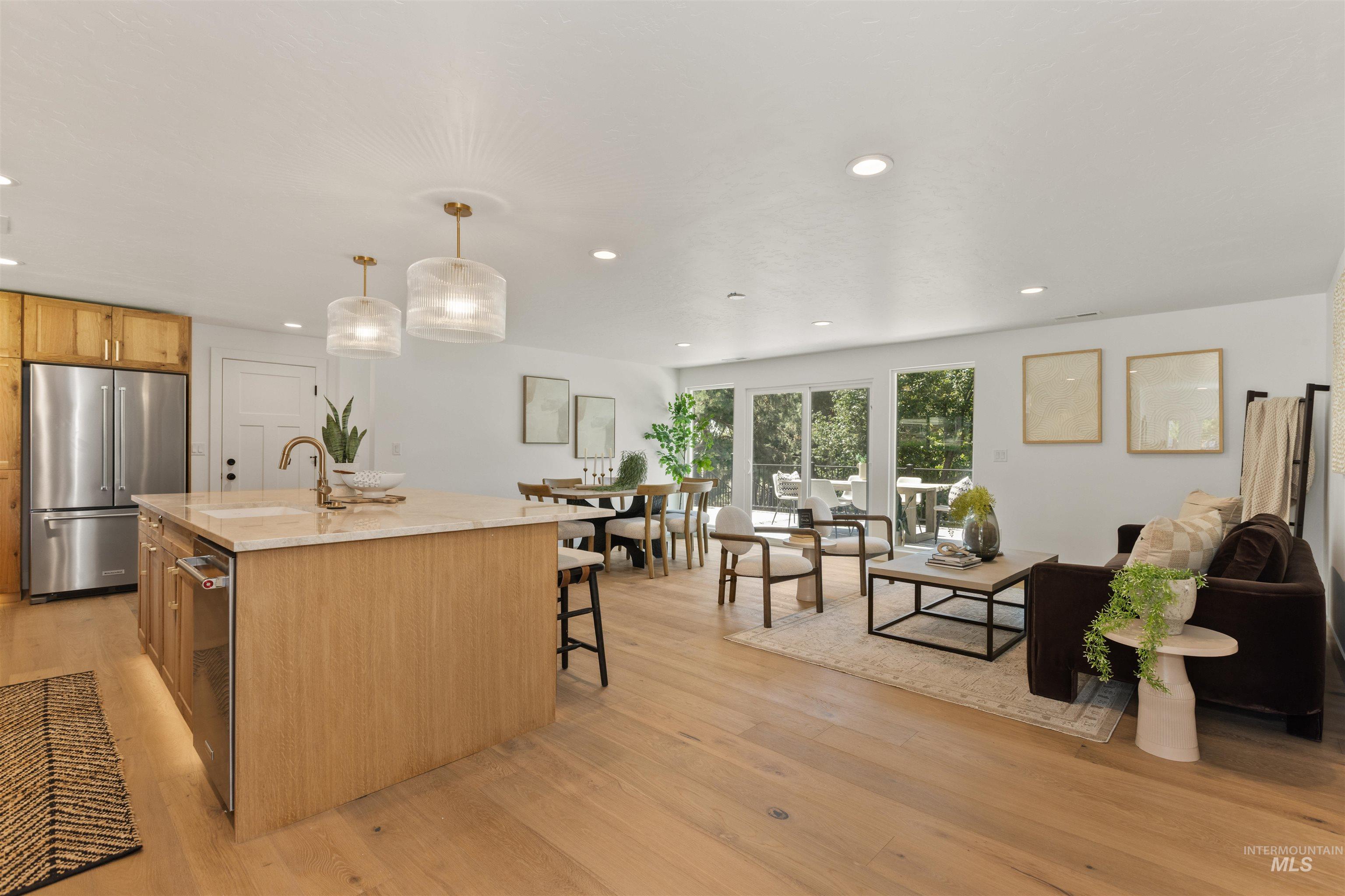 Kitchen with an island with sink, pendant lighting, light wood-type flooring, appliances with stainless steel finishes, and light stone counters