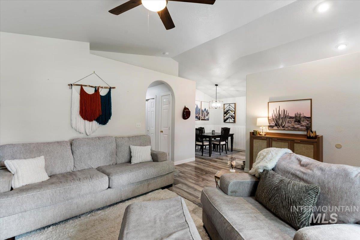 Living area featuring wood-finish flooring, vaulted ceilings, a ceiling fan with integrated lighting, and an arched doorway leading to a dining space with a chandelier
