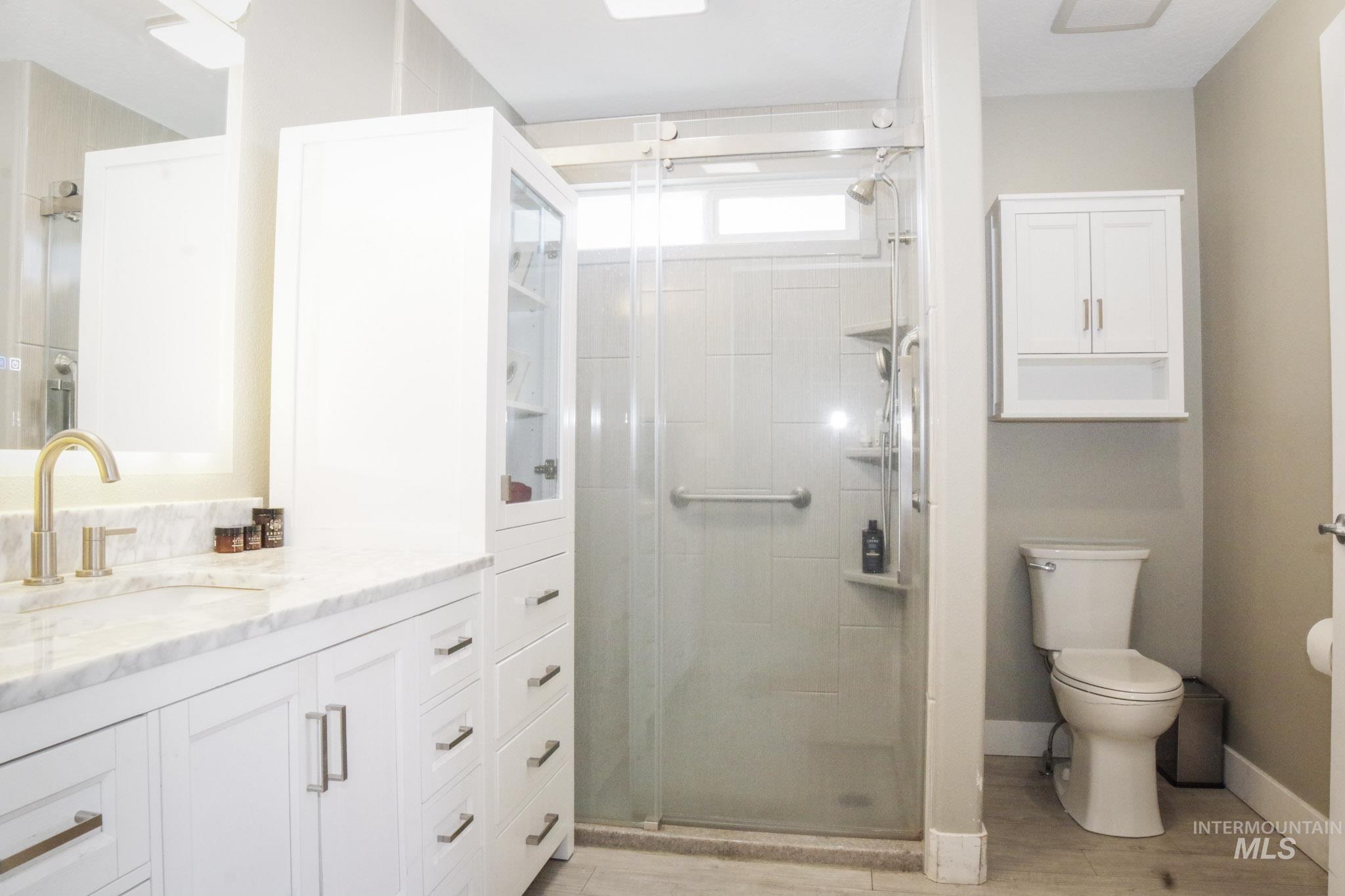Bathroom featuring a glass-enclosed shower with a transom window, a single vanity with a stone countertop, a built-in storage tower, and white shaker-style cabinetry with brushed nickel hardware