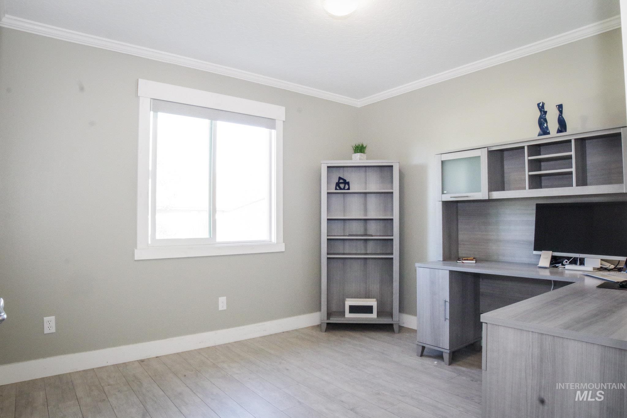 Light grey walls with white trim, wood-finish flooring, a built-in L-shaped desk with upper cabinetry, and a separate shelving unit