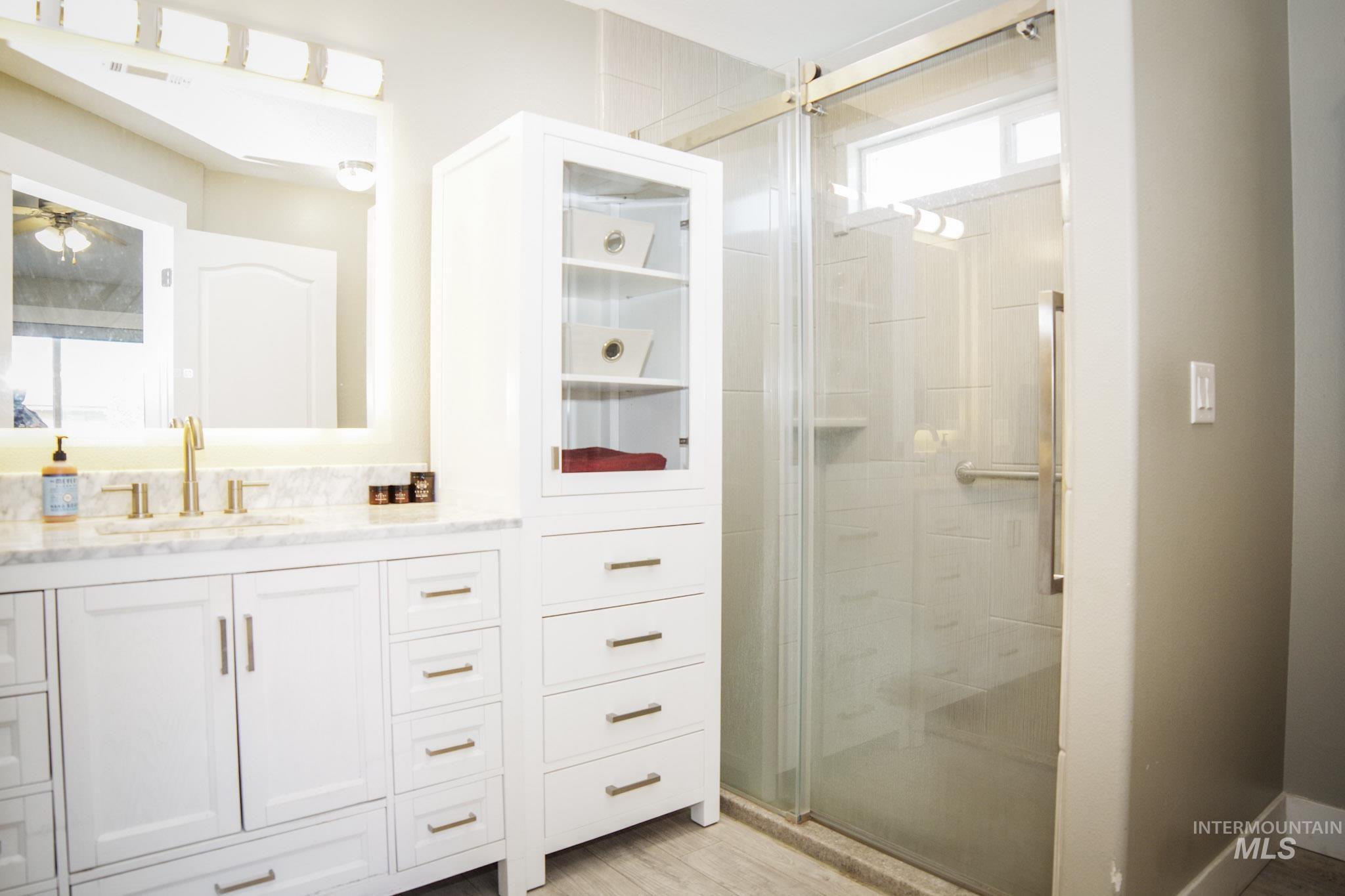 Bathroom featuring a white vanity with a rectangular sink and brushed gold hardware, a marble-finish countertop, a large illuminated mirror, a glass-enclosed shower, and a built-in storage cabinet