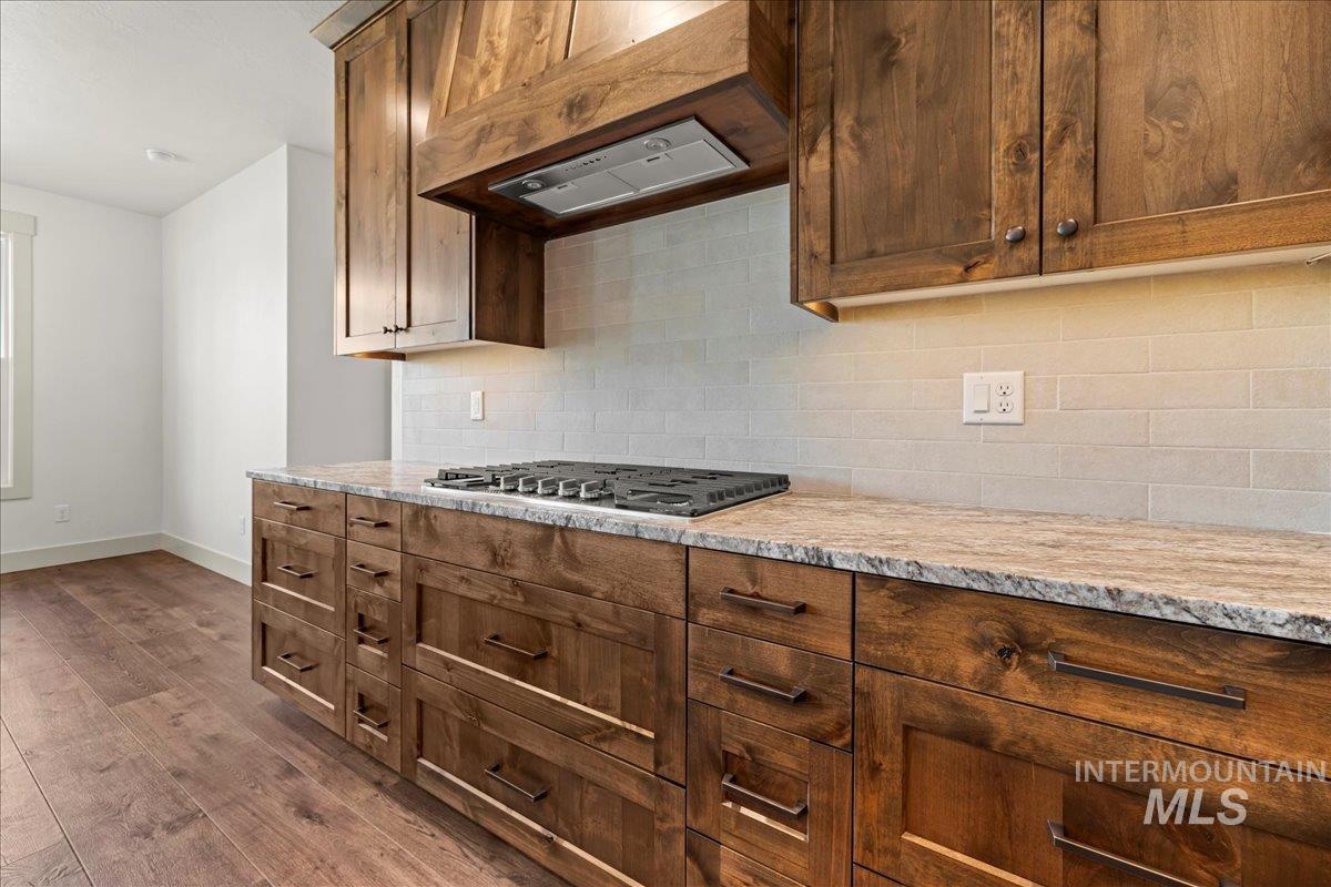 Kitchen featuring range hood, dark wood-type flooring, light stone countertops, decorative backsplash, and stainless steel gas stovetop