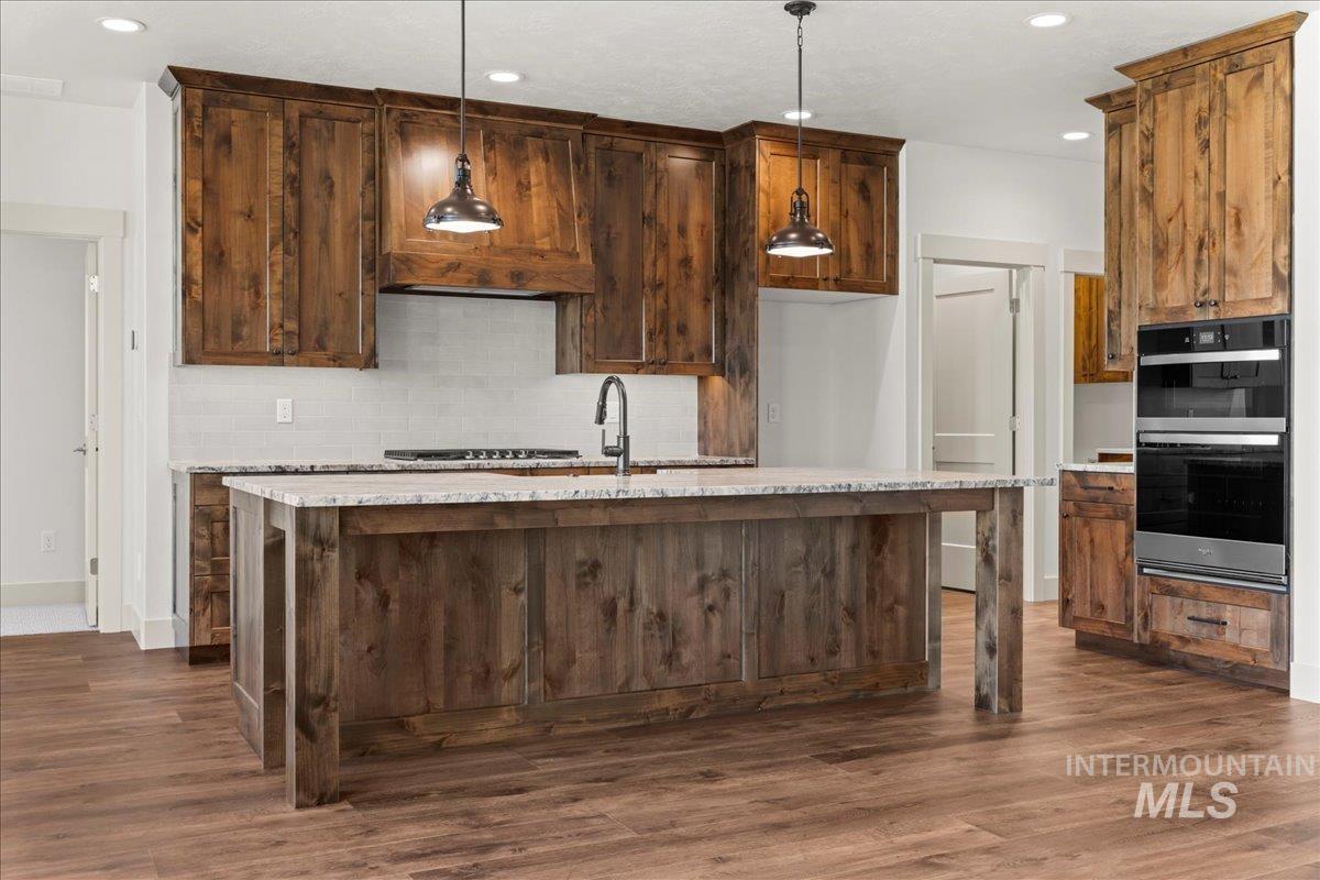 Kitchen featuring recessed lighting, dark wood-style floors, stainless steel double oven, decorative backsplash, and a center island with sink