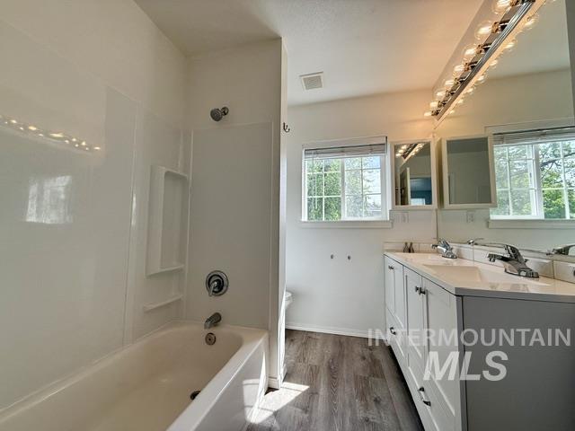 Bathroom featuring a white bathtub with integrated shower, dual vanity with white cabinetry, white countertops, two framed mirrors, and wood-finish flooring