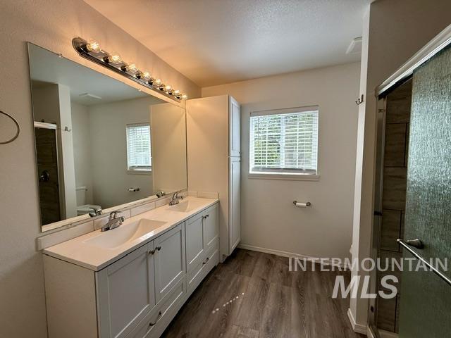 Bathroom featuring a dual vanity with white cabinetry and integrated sinks, a large mirror with a multi-bulb light fixture, wood-finish flooring, a window with blinds, and a glass-enclosed shower with tiled walls