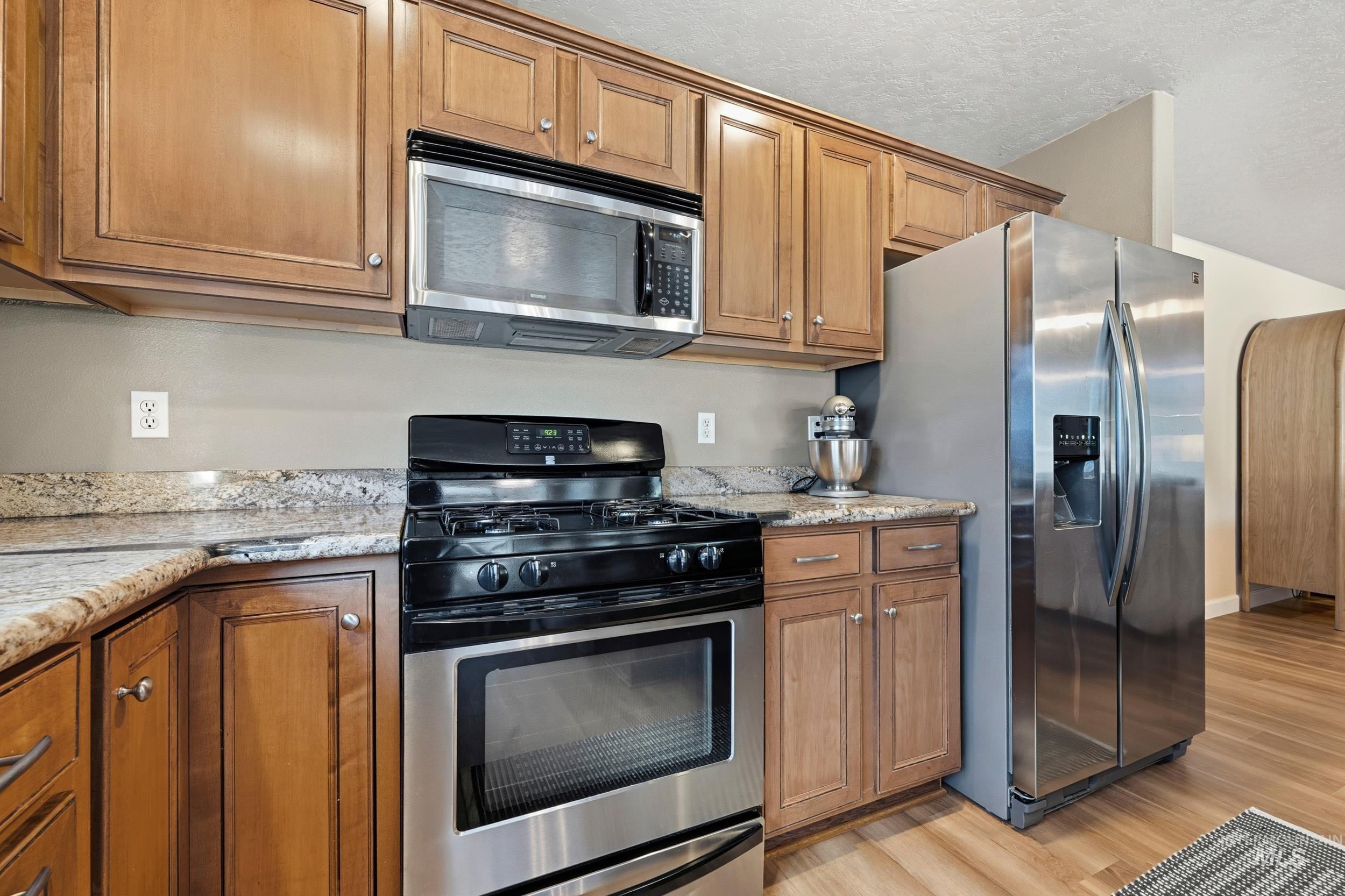 Kitchen with appliances with stainless steel finishes, brown cabinetry, light stone countertops, light wood-type flooring, and a textured ceiling