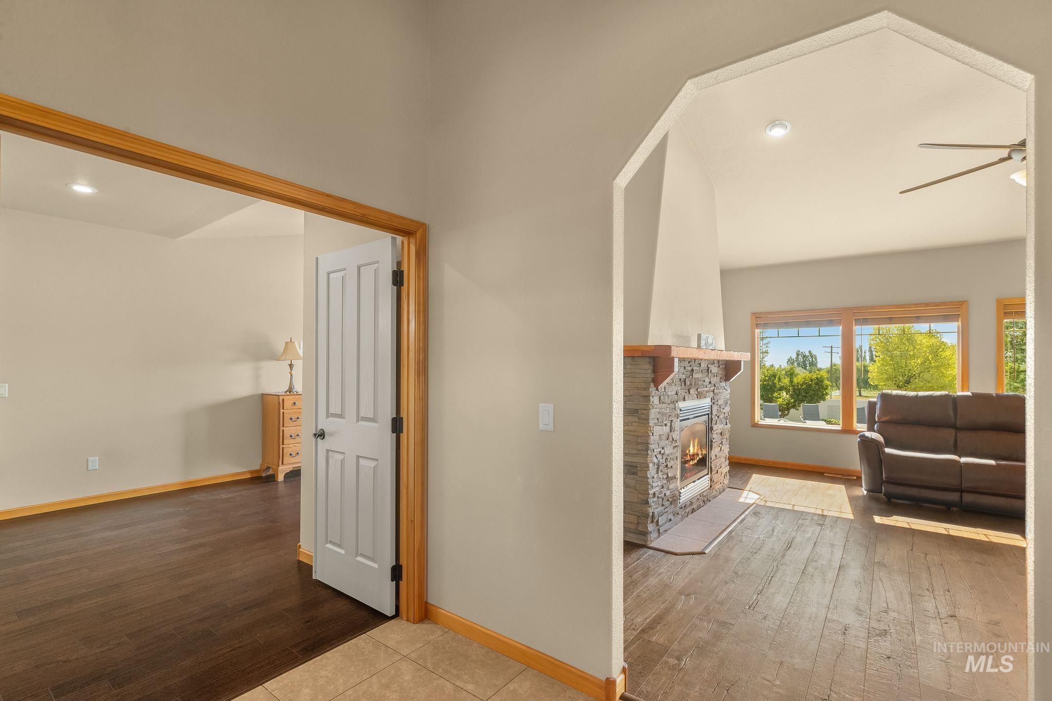Entryway with tile flooring opening to a living area featuring a stone-clad fireplace with a wood mantle, wood-finish flooring, large windows, and a ceiling fan