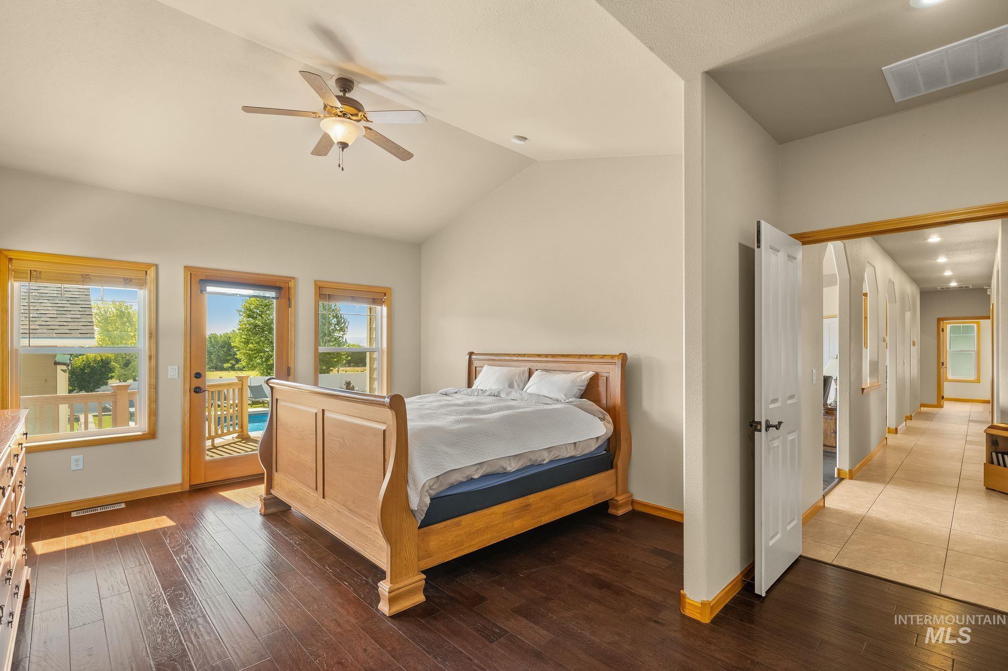 Vaulted ceiling bedroom with wood-finish flooring, featuring a ceiling fan, direct access to an outdoor deck, and expansive windows
