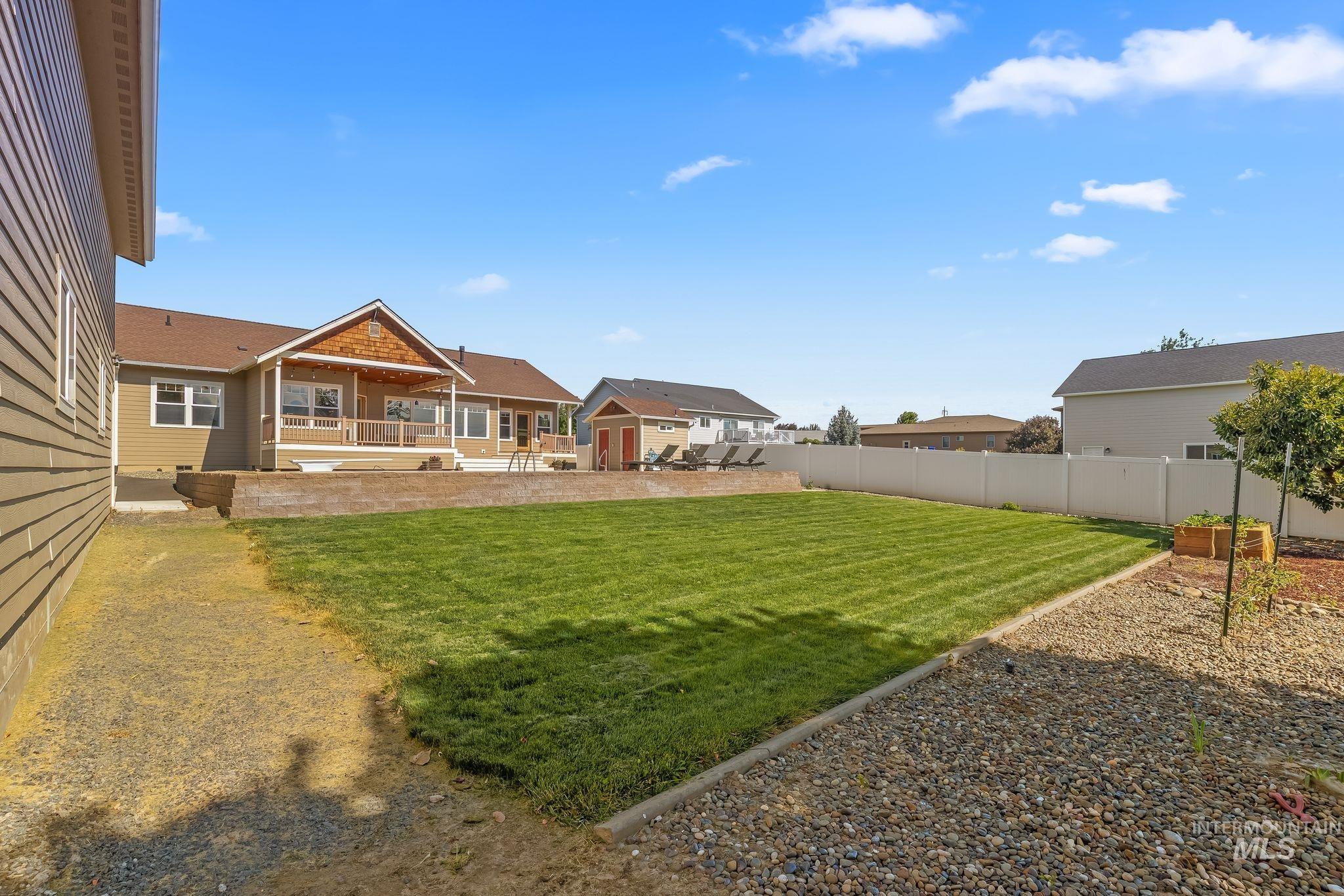 Expansive backyard featuring a large manicured lawn, gravel landscaping, a white privacy fence, a raised patio, and an outdoor shed