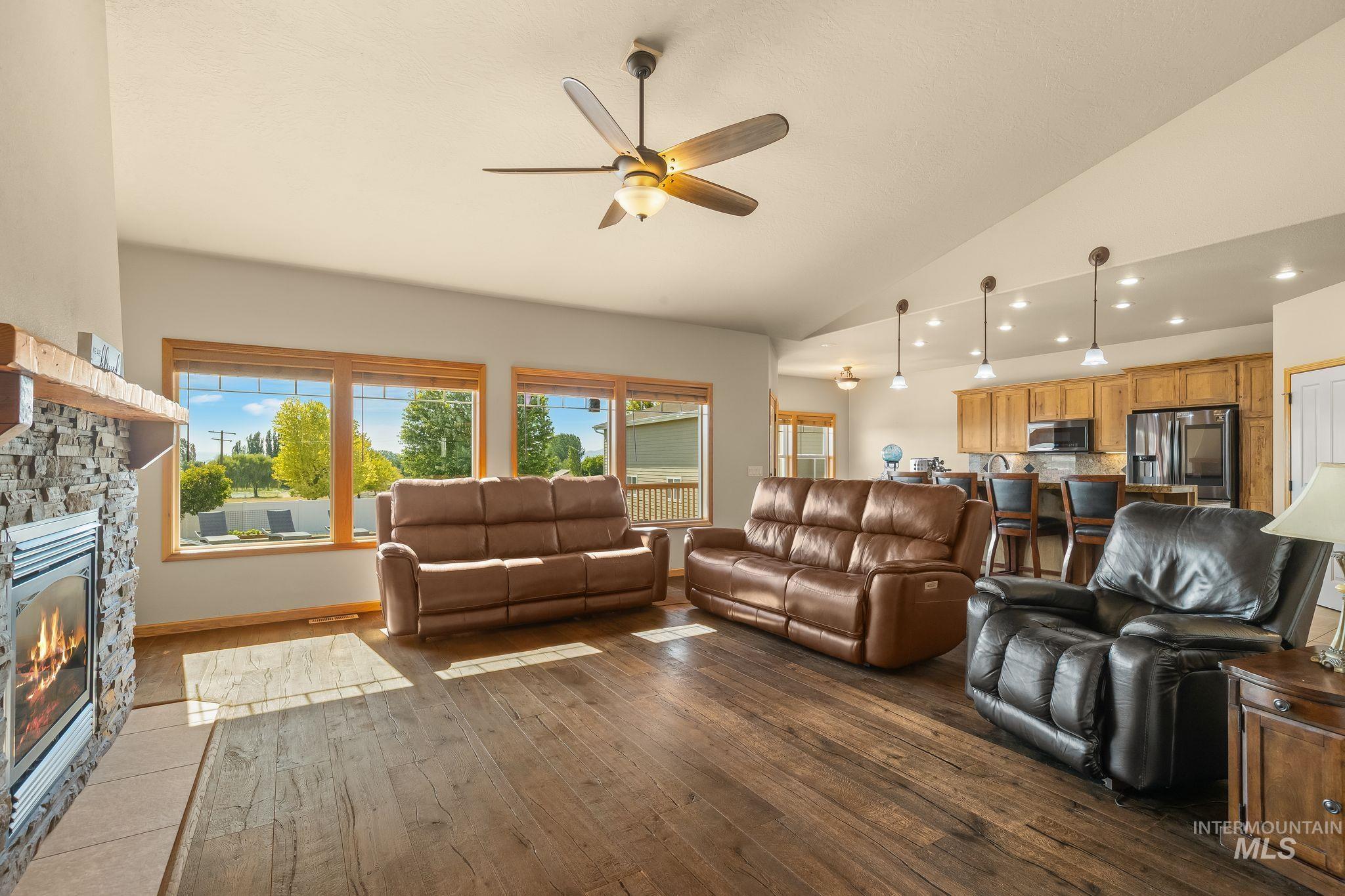 Vaulted ceiling living area featuring wood-finish flooring, a stone-faced fireplace with a wood mantle, and large windows with wood trim