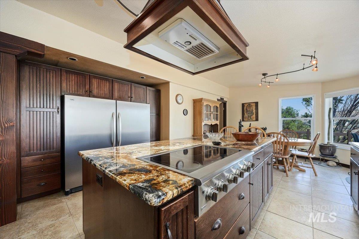 Kitchen featuring range hood, appliances with stainless steel finishes, an island with sink, dark brown cabinets, and light stone countertops