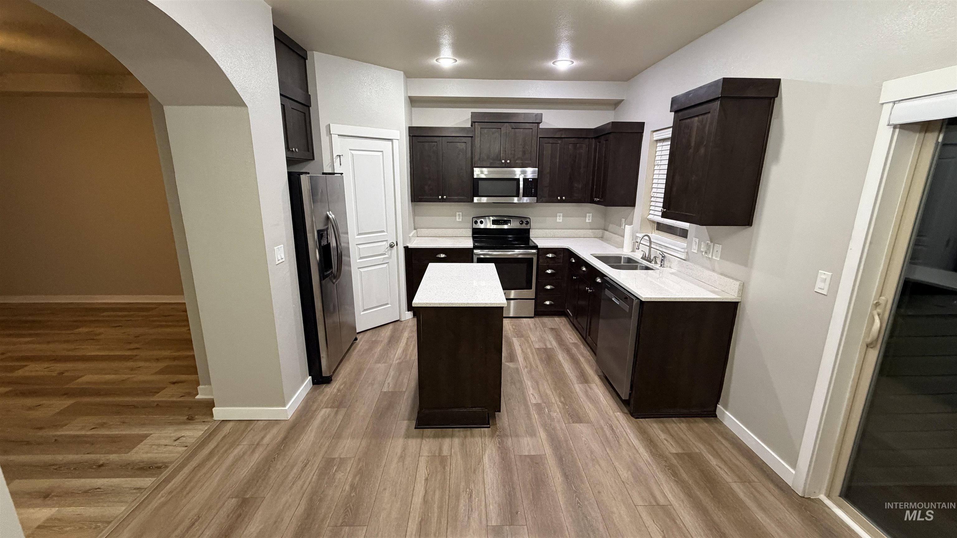 Modern kitchen featuring dark wood cabinetry, stainless steel appliances, light countertops, wood-finish flooring, and a central island