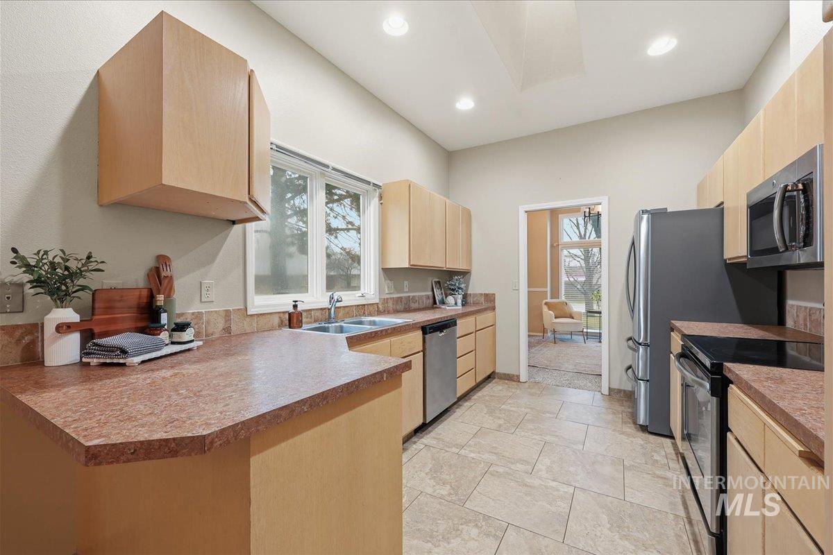 Kitchen with light brown cabinetry, appliances with stainless steel finishes, a peninsula, recessed lighting, and stone tile flooring