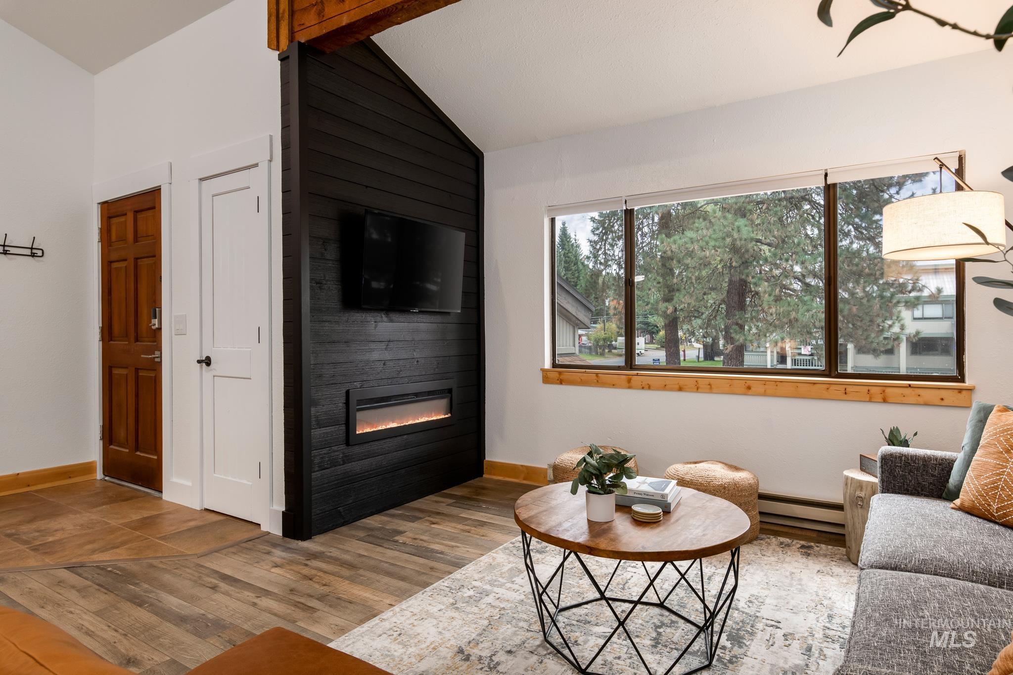 Living area featuring a floor-to-ceiling accent wall with a built-in linear electric fireplace, wood-finish flooring, and exposed ceiling beams